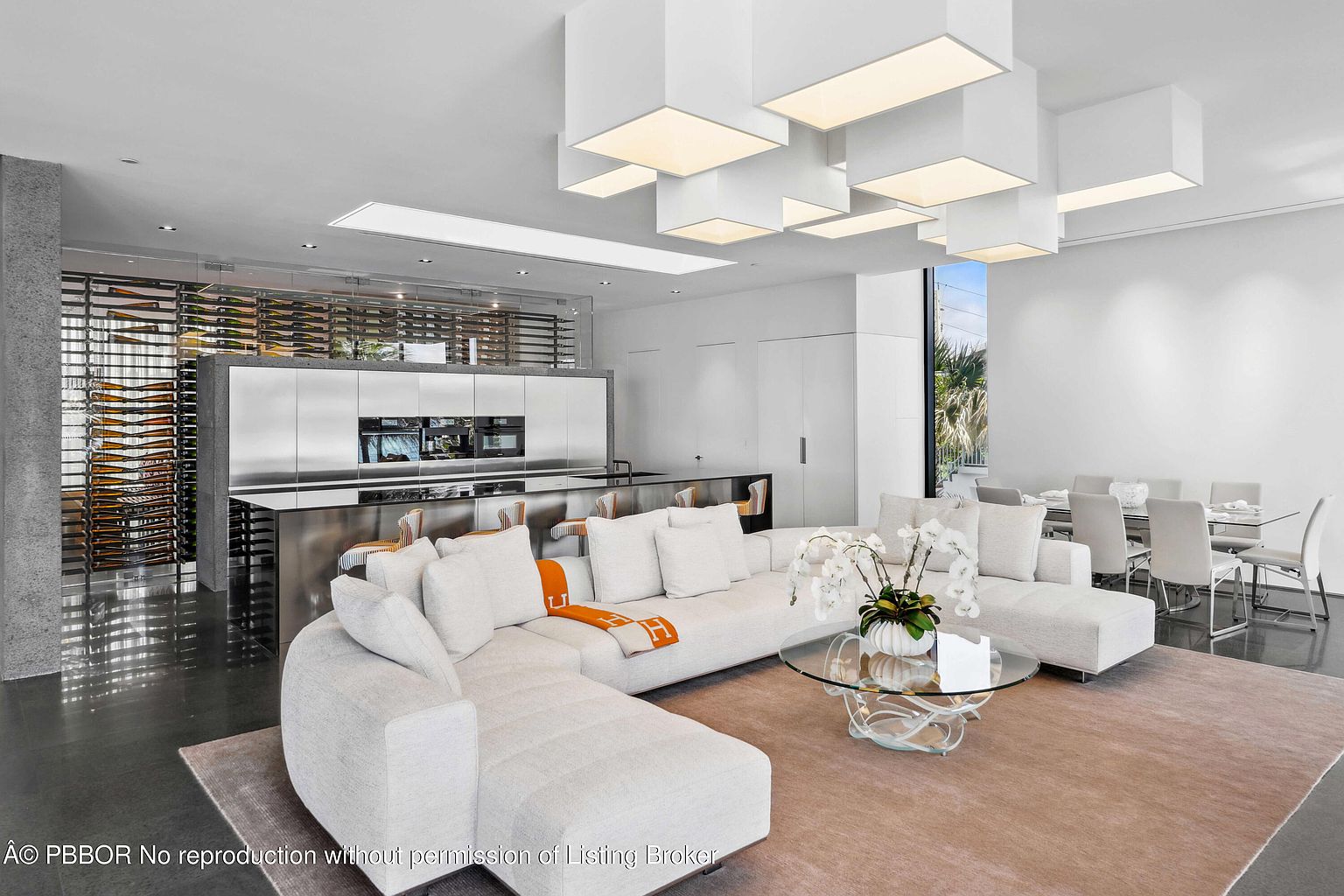 This is a bright and modern living room featuring a large, white sectional sofa, a unique glass coffee table, and a custom lighting fixture with multiple square lights. The open-concept design seamlessly integrates the living area with the kitchen, which includes a wine rack and stainless steel appliances. The overall impression is one of luxury and contemporary elegance.