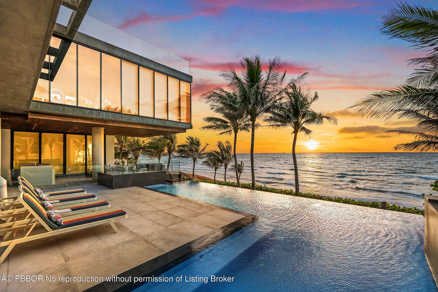 This image showcases a luxurious infinity pool overlooking the ocean at sunset. The modern architecture of the adjacent building features large glass windows, reflecting the warm light. Palm trees line the coast, adding to the tropical ambiance and highlighting the property's prime beachfront location.