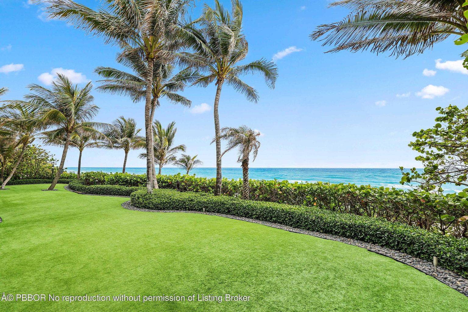 This exterior shot showcases a meticulously maintained yard with lush green grass and a variety of palm trees. A neatly trimmed hedge borders the lawn, providing a clear view of the ocean in the background. The scene evokes a sense of tranquility and luxury, highlighting the property's desirable location and outdoor space.