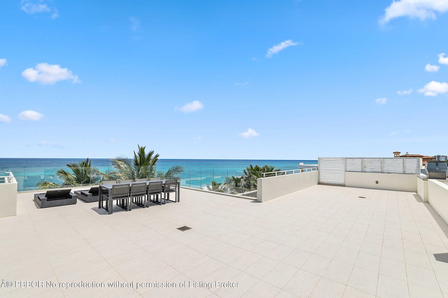 This image showcases a spacious rooftop patio or deck with a stunning ocean view. The area is furnished with modern outdoor furniture, including a dining set and lounge seating. The deck features light-colored tiling and a glass railing, enhancing the sense of openness and luxury, while palm trees add a tropical touch.