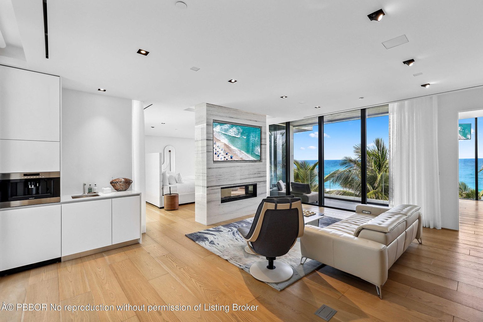 This is a bright and modern living room with floor-to-ceiling windows offering ocean views. The room features a neutral color palette, light wood flooring, and contemporary furniture including a sleek sofa and swivel chair. A marble-clad fireplace and TV wall add a luxurious touch, while the open layout creates a spacious and airy feel.