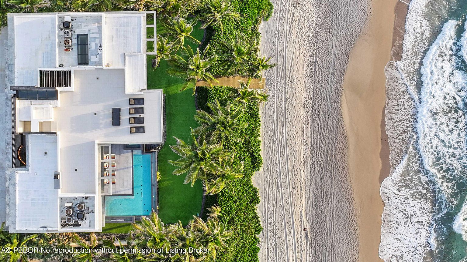 This aerial shot showcases a luxurious beachfront property with a modern architectural design. The house features a flat white roof, a swimming pool, and a well-manicured lawn that transitions into lush tropical vegetation bordering a pristine sandy beach. The ocean waves gently crash onto the shore, creating a serene and exclusive atmosphere.