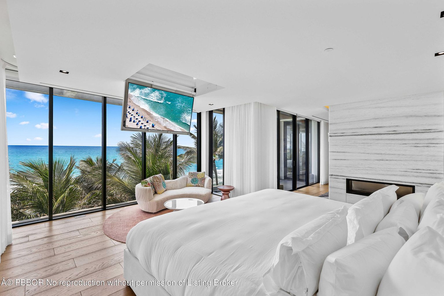 This is a luxurious primary bedroom featuring a large bed with white linens, floor-to-ceiling windows offering ocean views, and a modern fireplace with marble surround. A curved sofa and a pop-up television add to the room's elegance and functionality. The overall impression is one of serene opulence and comfort.