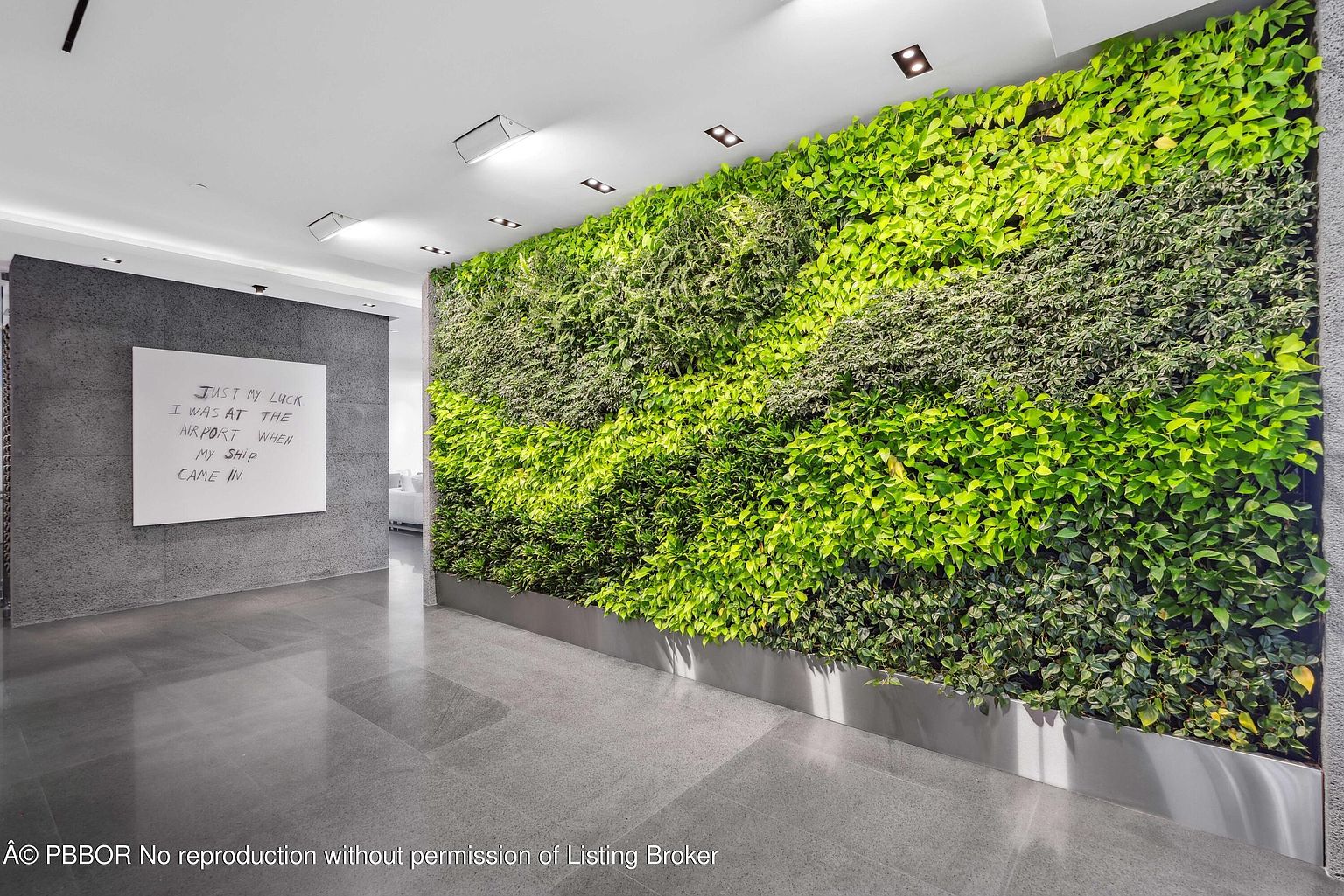 This interior shot showcases a modern hallway featuring a striking green wall, adding a touch of nature to the space. A large, framed text art piece hangs on the adjacent gray wall, creating a contemporary and artistic atmosphere. The polished floors reflect the light, enhancing the spacious feel of the hallway.