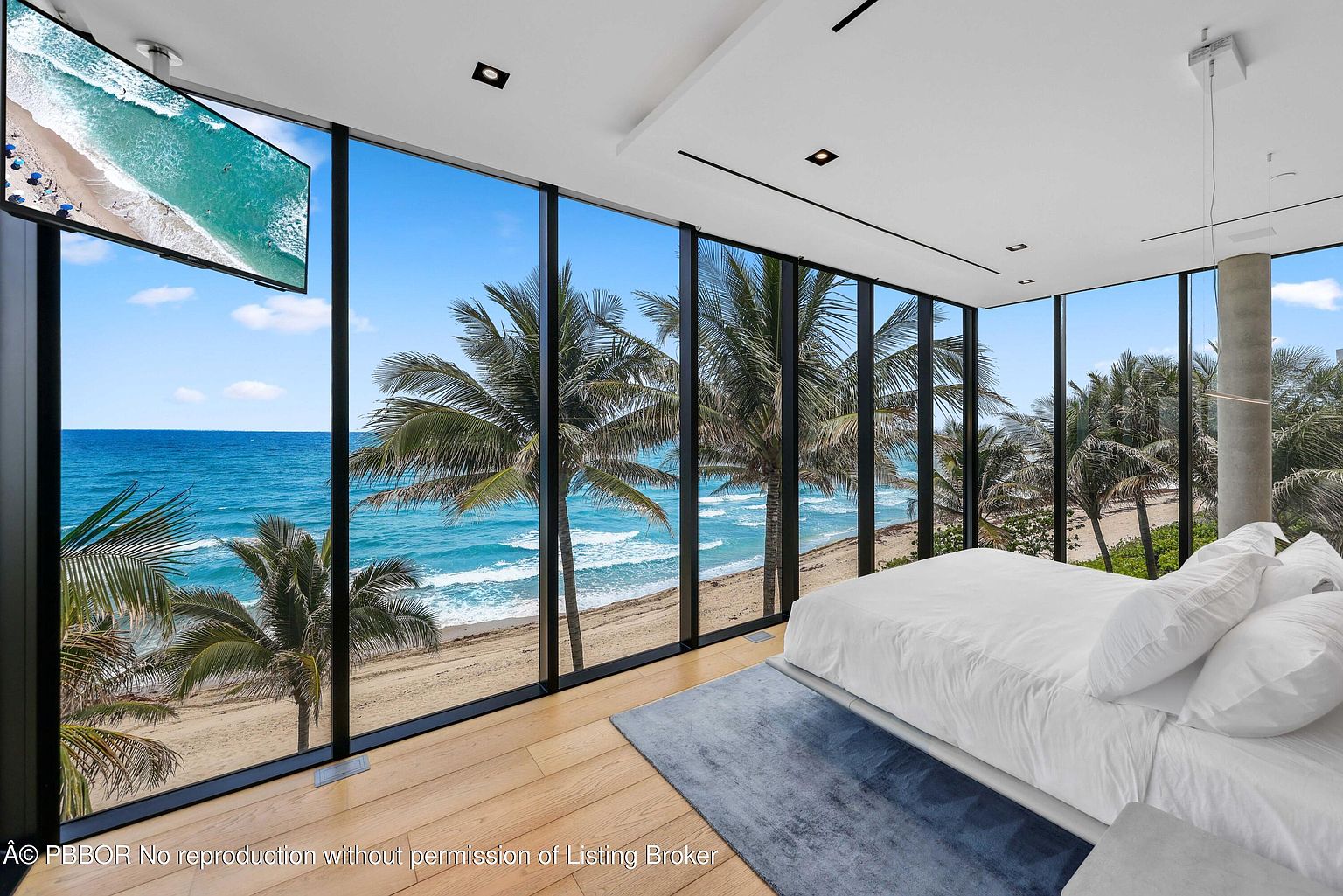 This is a luxurious primary bedroom with floor-to-ceiling windows offering stunning ocean views framed by palm trees. The room features a minimalist design with a large bed, a blue rug, and a wall-mounted television. The overall impression is one of tranquility and high-end coastal living.