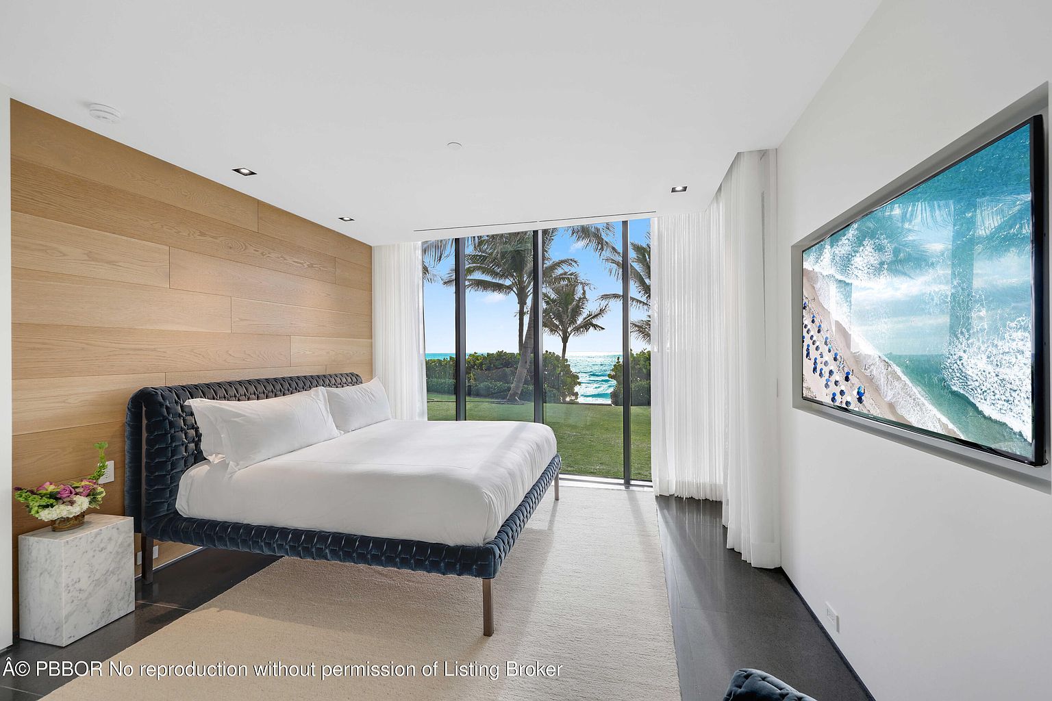 This is a luxurious primary bedroom featuring a large bed with a tufted headboard and crisp white linens. A wall of windows offers stunning views of palm trees and the ocean, while a modern TV is mounted on the opposite wall. The room is decorated in a minimalist style with a wood accent wall and a neutral color palette, creating a serene and inviting atmosphere.