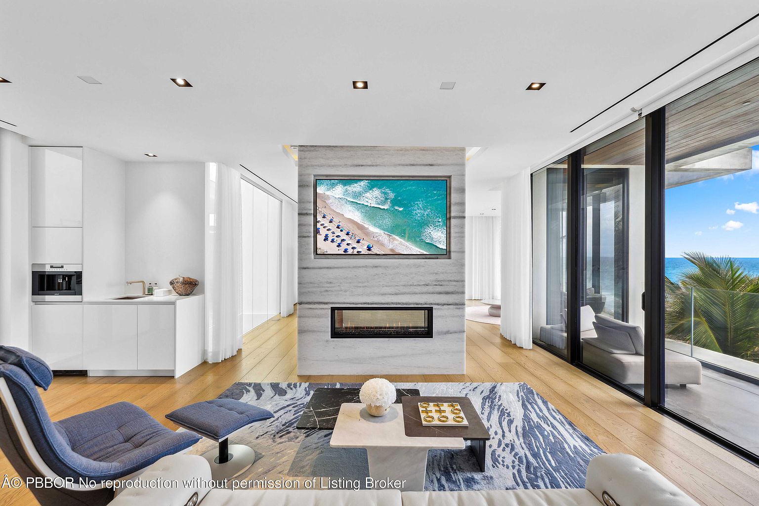 This is a bright and modern living room featuring a marble fireplace with a large television above, floor-to-ceiling windows offering ocean views, and contemporary furniture. The open floor plan and light wood flooring create a spacious and airy feel, while the blue and white area rug adds a touch of color and texture. The room exudes luxury and sophistication.