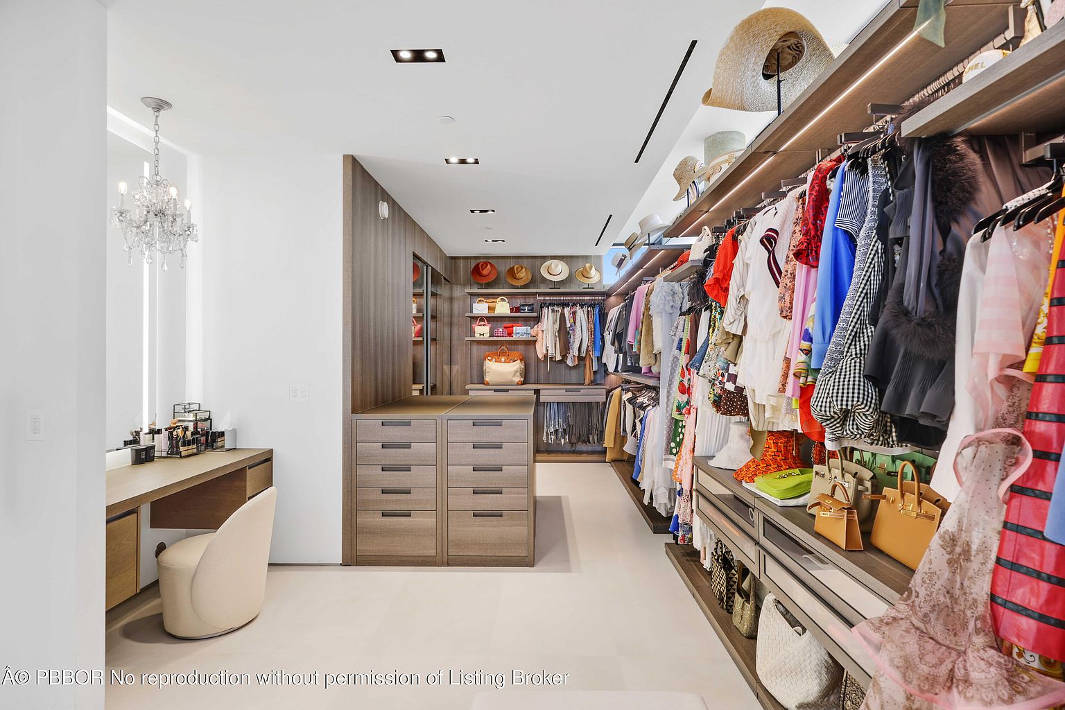 This is a well-organized walk-in closet featuring custom wood cabinetry with drawers and shelving. The closet is filled with a variety of clothing, handbags, and hats, showcasing ample storage space. A vanity area with a chandelier adds a touch of luxury to the space.