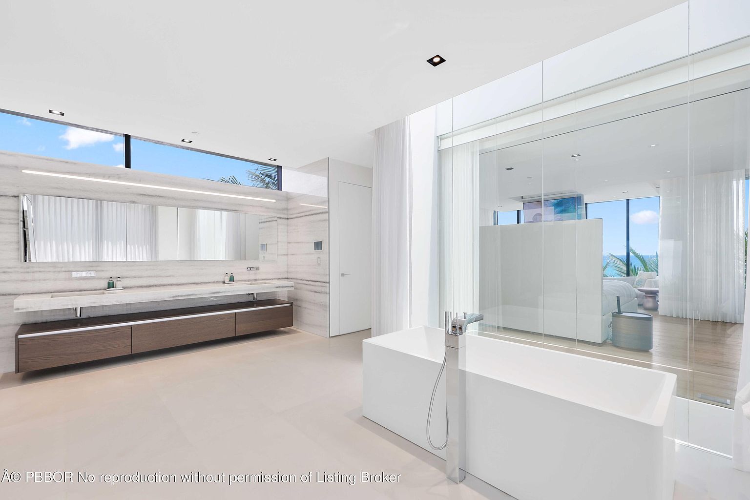 This is a luxurious primary bathroom featuring a modern freestanding bathtub, a large vanity with dual sinks, and a spacious walk-in shower enclosed in glass. The color palette is predominantly white and neutral, creating a clean and serene atmosphere. The bathroom also offers a view of the ocean, enhancing the spa-like experience.