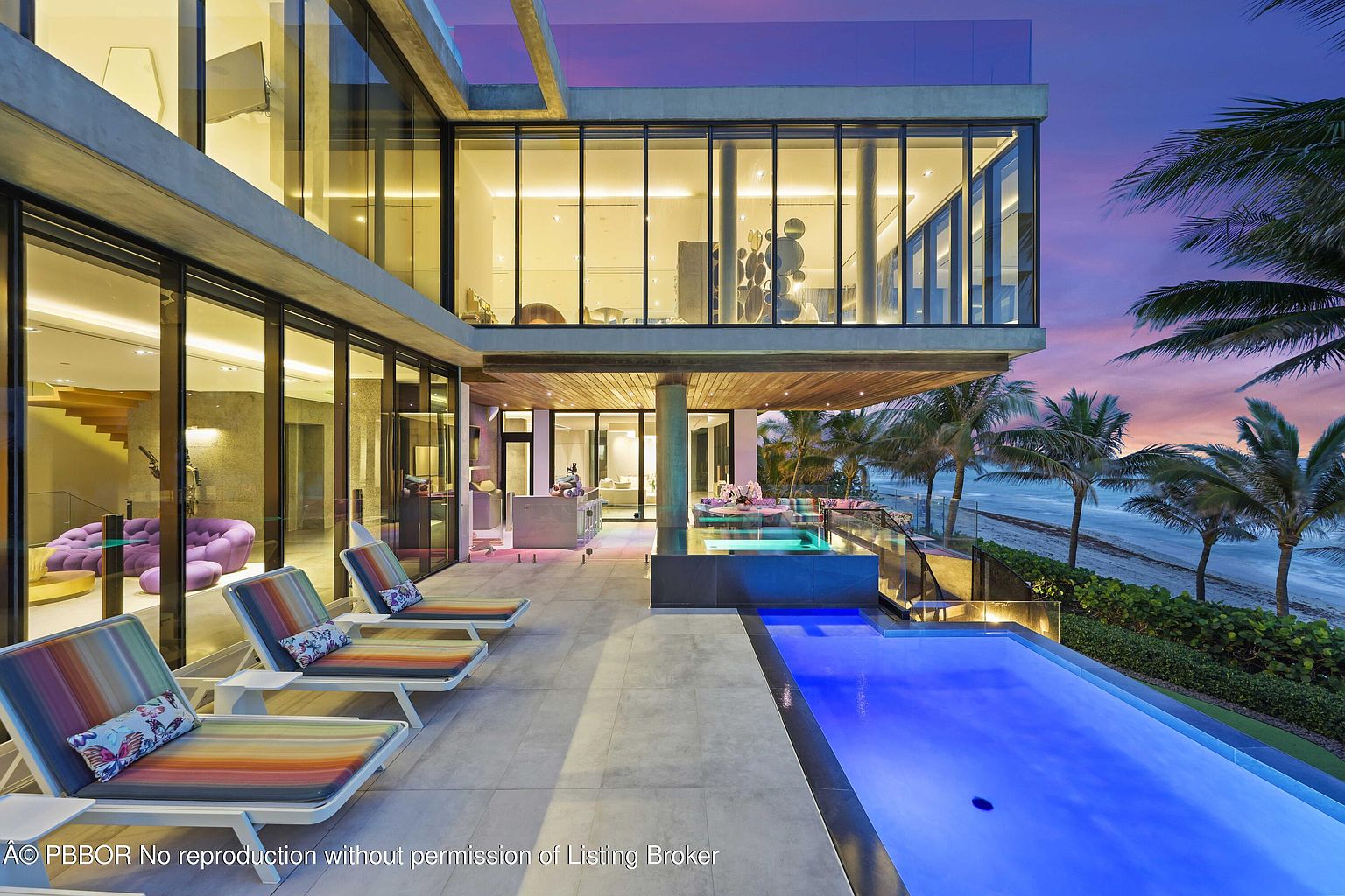 This image showcases a luxurious modern home's exterior, focusing on the pool and patio area. The infinity pool extends towards the beach, framed by palm trees and lush greenery. The house features large glass windows, offering stunning views and creating a seamless indoor-outdoor living experience, with lounge chairs positioned for relaxation by the pool.