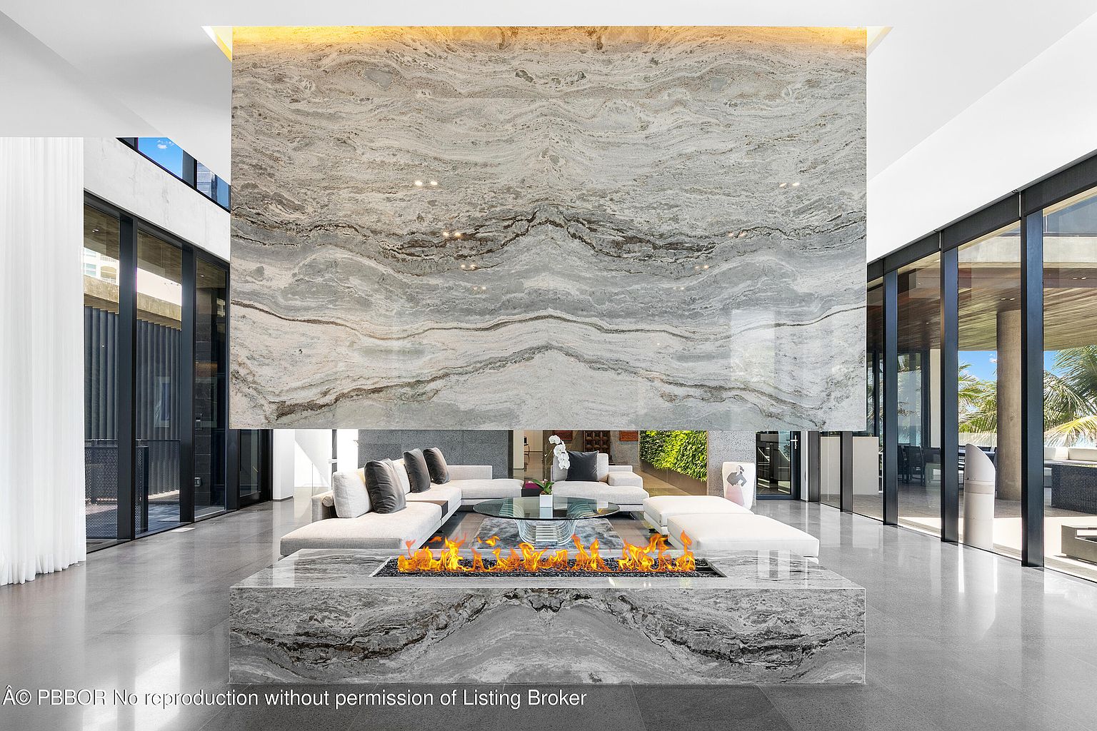 This is a luxurious living room featuring a modern fireplace with a marble surround and base, creating a focal point. The room is furnished with white sectional sofas and a glass coffee table, complemented by large windows that offer ample natural light. The overall design is sleek and contemporary, emphasizing spaciousness and high-end finishes.