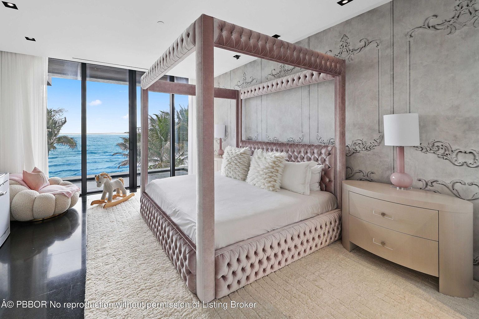 This is a luxurious bedroom featuring a four-poster bed with tufted upholstery in a soft pink hue. The room is decorated with a neutral color palette, including a light-colored rug and elegant wallpaper with ornate details. Large windows offer a view of the ocean, creating a serene and upscale atmosphere.