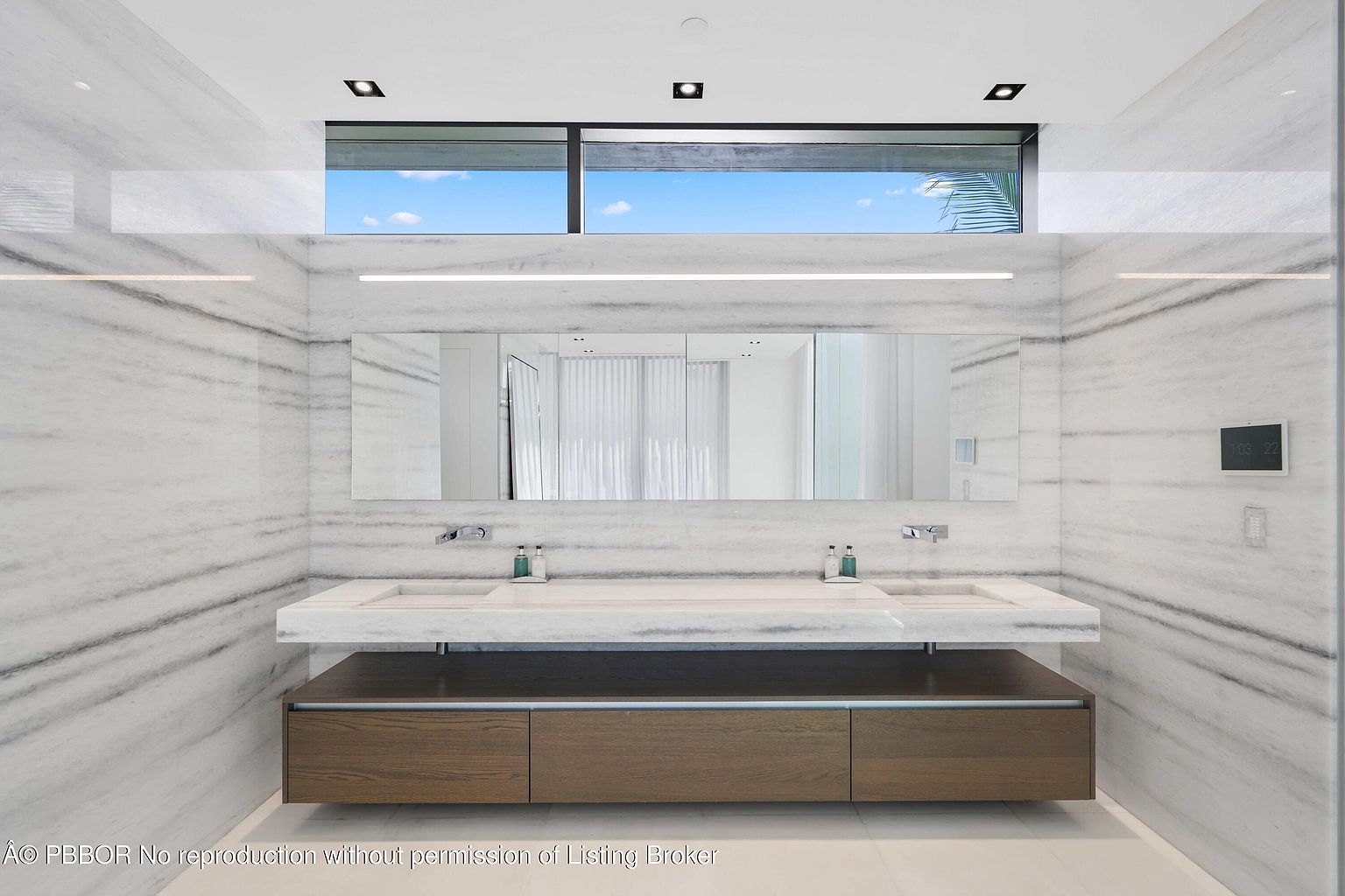 This is a modern primary bathroom featuring a double vanity with integrated sinks and a sleek, minimalist design. The walls are clad in marble, and a long, rectangular mirror spans the length of the vanity. A horizontal window above the mirror provides natural light, and the overall aesthetic is clean, luxurious, and spa-like.