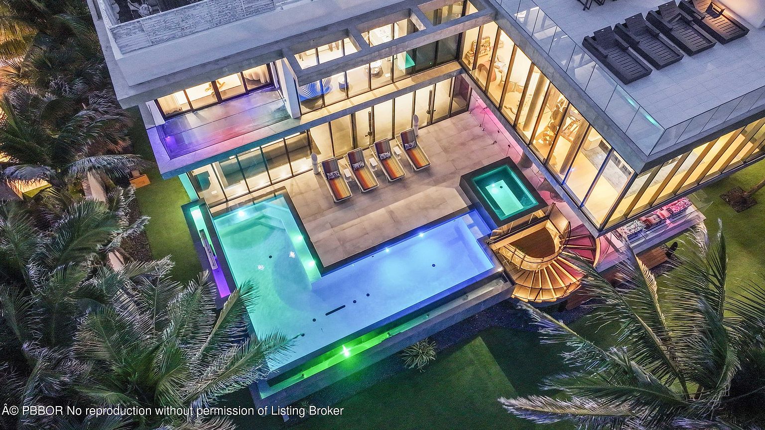 This aerial view showcases a modern luxury home with a stunning pool area featuring multiple pools and a jacuzzi. The property boasts expansive glass walls, a spiral staircase, and a rooftop deck with lounge chairs. Lush landscaping surrounds the home, creating a private and serene oasis.