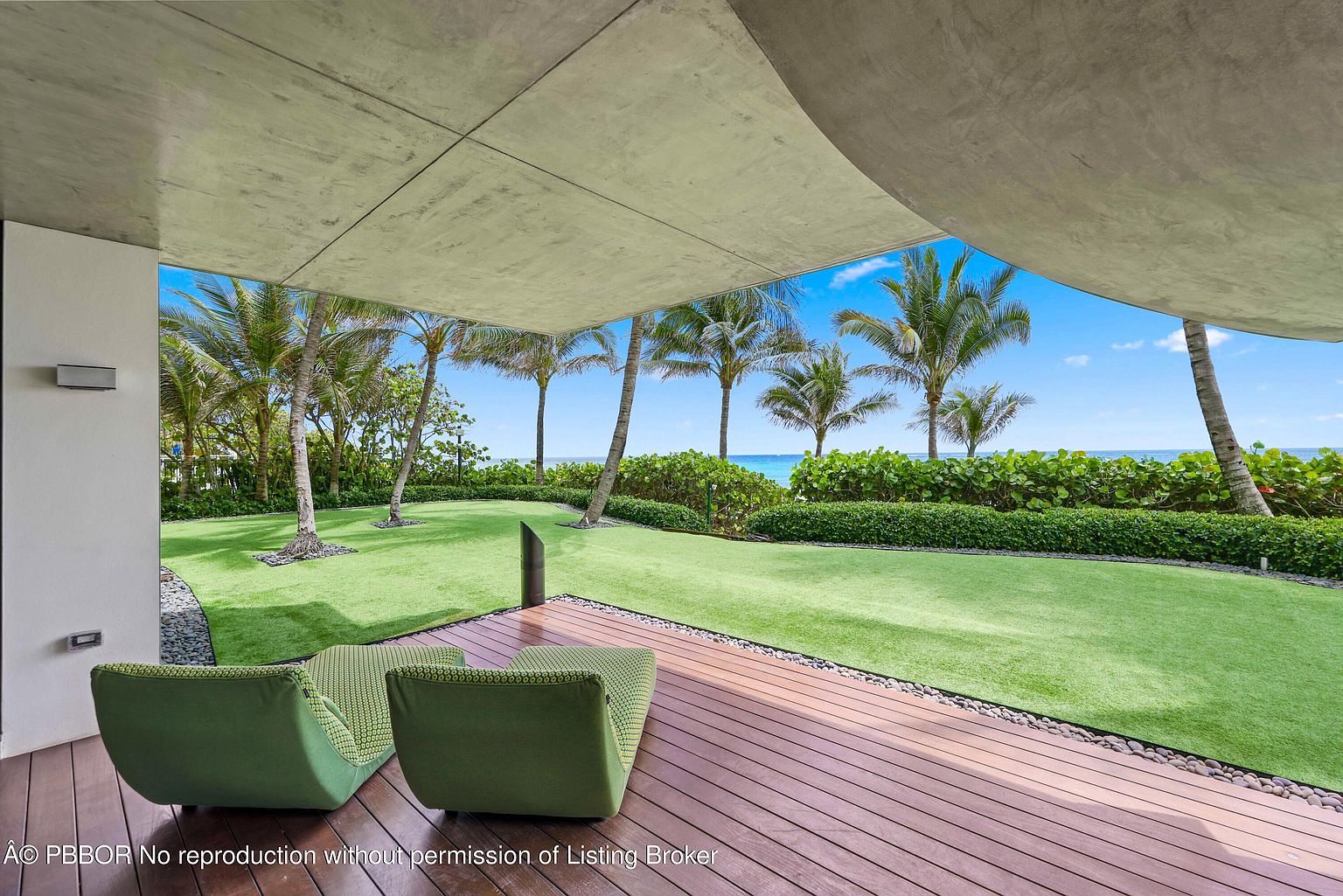This image showcases a luxurious outdoor patio area with a modern design. The patio features a wooden deck with comfortable green seating, overlooking a lush green lawn and palm trees leading to the ocean. The concrete overhang provides shade and architectural interest, creating a serene and inviting outdoor living space.