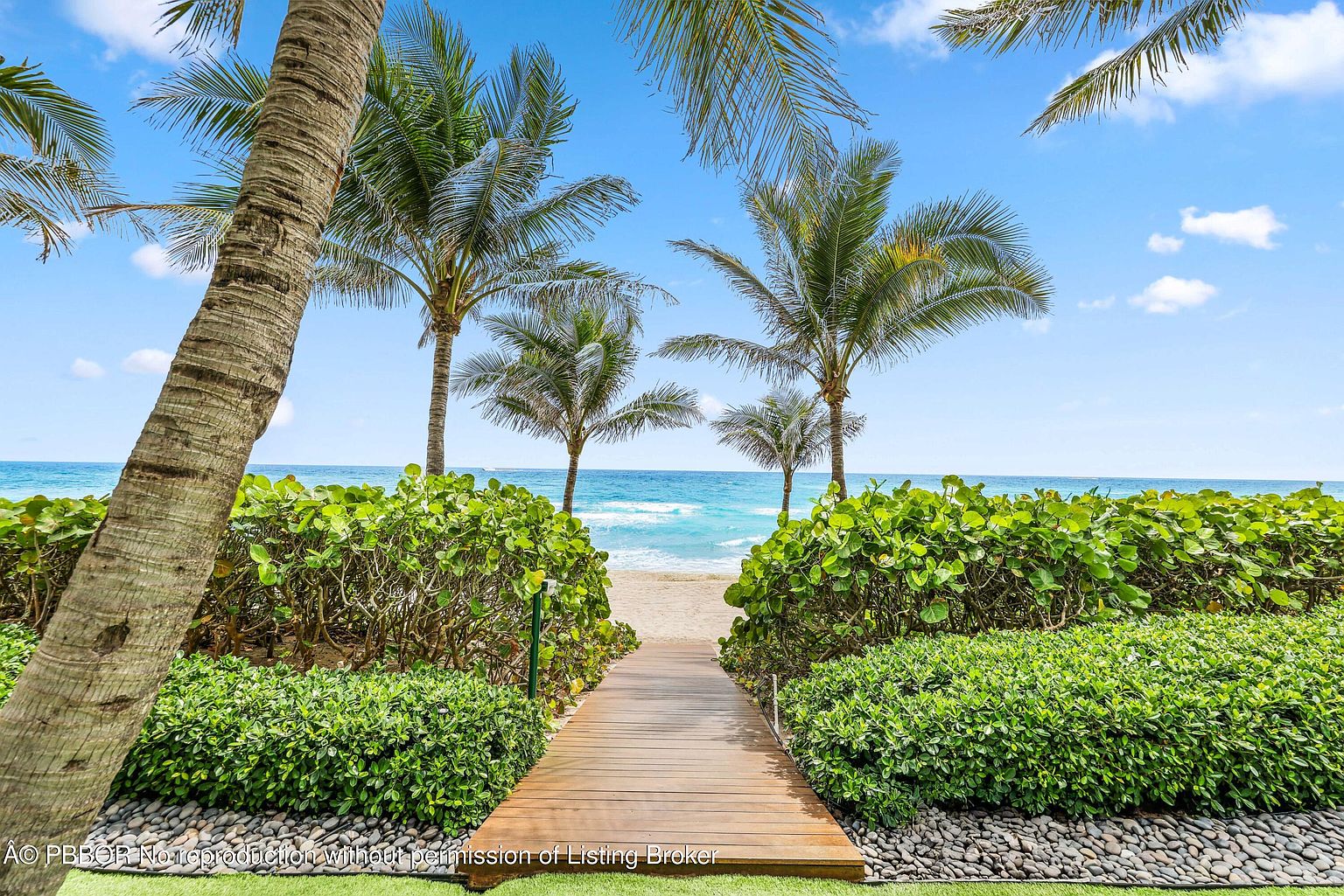 This inviting exterior shot showcases a wooden boardwalk leading to a pristine beach, framed by lush greenery and swaying palm trees. The turquoise ocean and clear blue sky create a serene and tropical atmosphere, perfect for highlighting the property's access to a private beach or waterfront amenity. The perspective is from the entrance of the boardwalk, inviting the viewer to walk towards the beach.