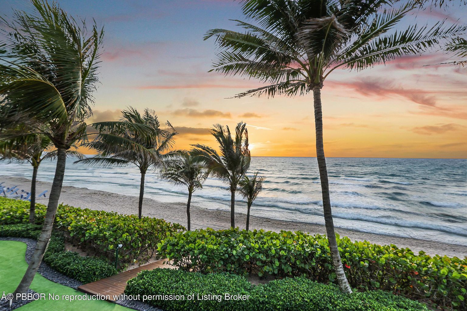 This image showcases a beautiful beachfront yard with lush green landscaping and several palm trees. A wooden walkway leads from the manicured garden area towards the sandy beach and ocean. The scene is bathed in the warm light of a sunset, creating a serene and inviting atmosphere.
