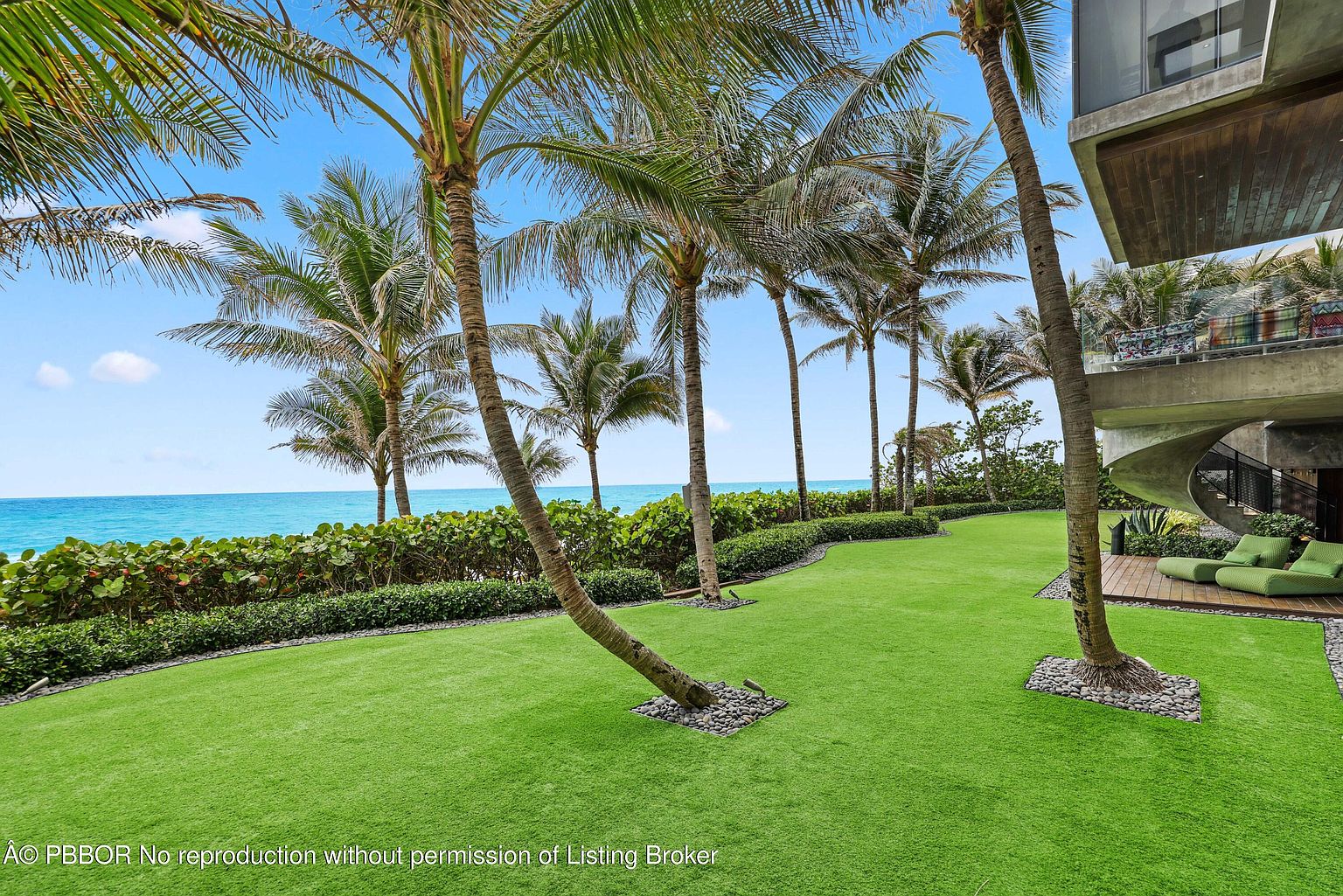 This image showcases a beautifully landscaped yard with lush green grass and several palm trees, creating a tropical ambiance. The yard extends towards the ocean, visible in the background, and a modern building with a balcony is partially visible on the right. The scene evokes a sense of luxury and relaxation, highlighting the property's outdoor space and proximity to the beach.
