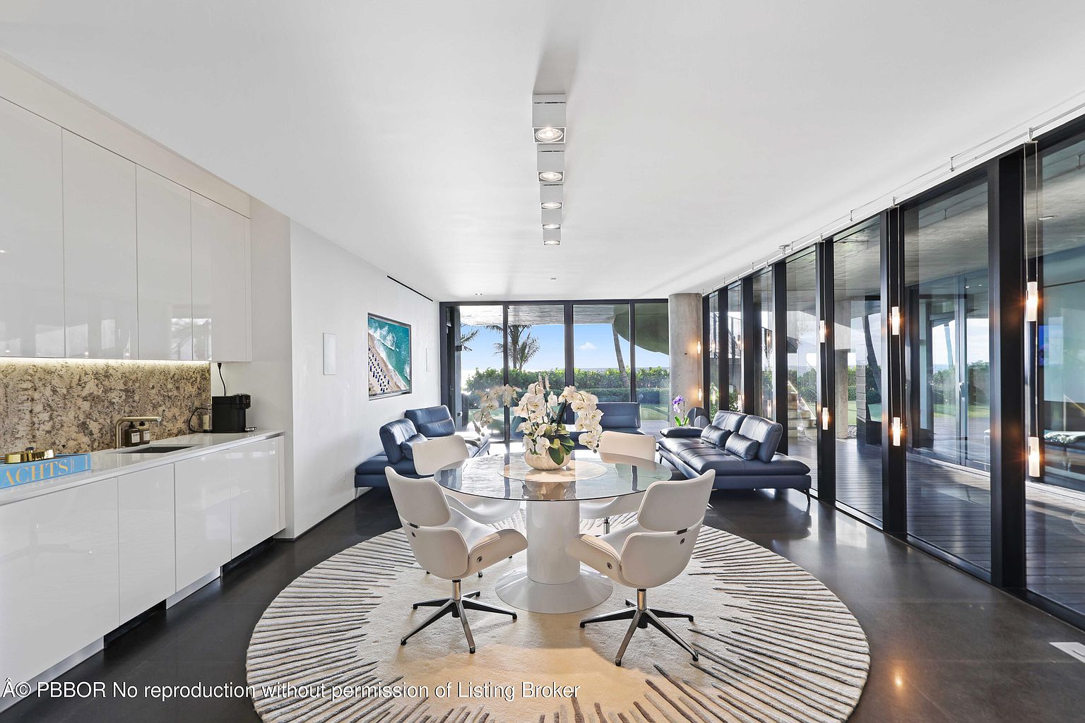 This interior shot showcases a modern dining area with a round glass table surrounded by four white chairs on a circular rug. The room features floor-to-ceiling windows offering an outdoor view, and is flanked by blue sofas, creating a luxurious and open living space. The perspective is from a medium distance, capturing the entire dining set and its integration with the surrounding living area.
