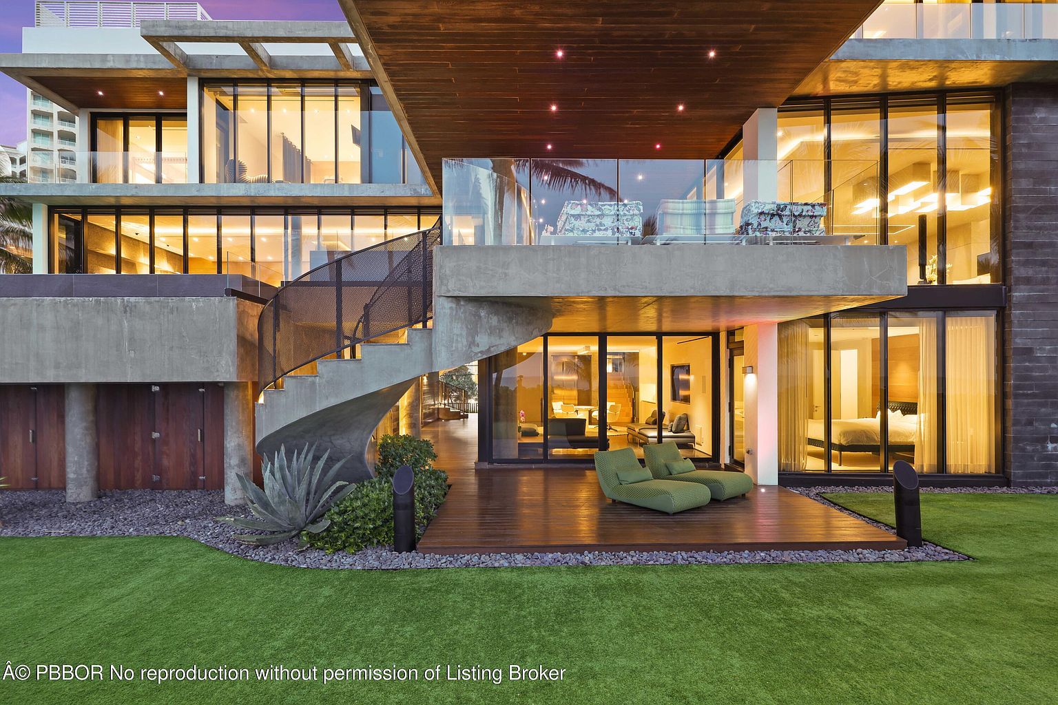 This is a rear view of a modern, multi-level home featuring expansive glass windows and doors, a concrete and wood exterior, and a spiral staircase. The property includes a wooden deck with lounge seating, manicured lawn, and lush landscaping. The architecture emphasizes clean lines and open spaces, creating a luxurious and inviting atmosphere.