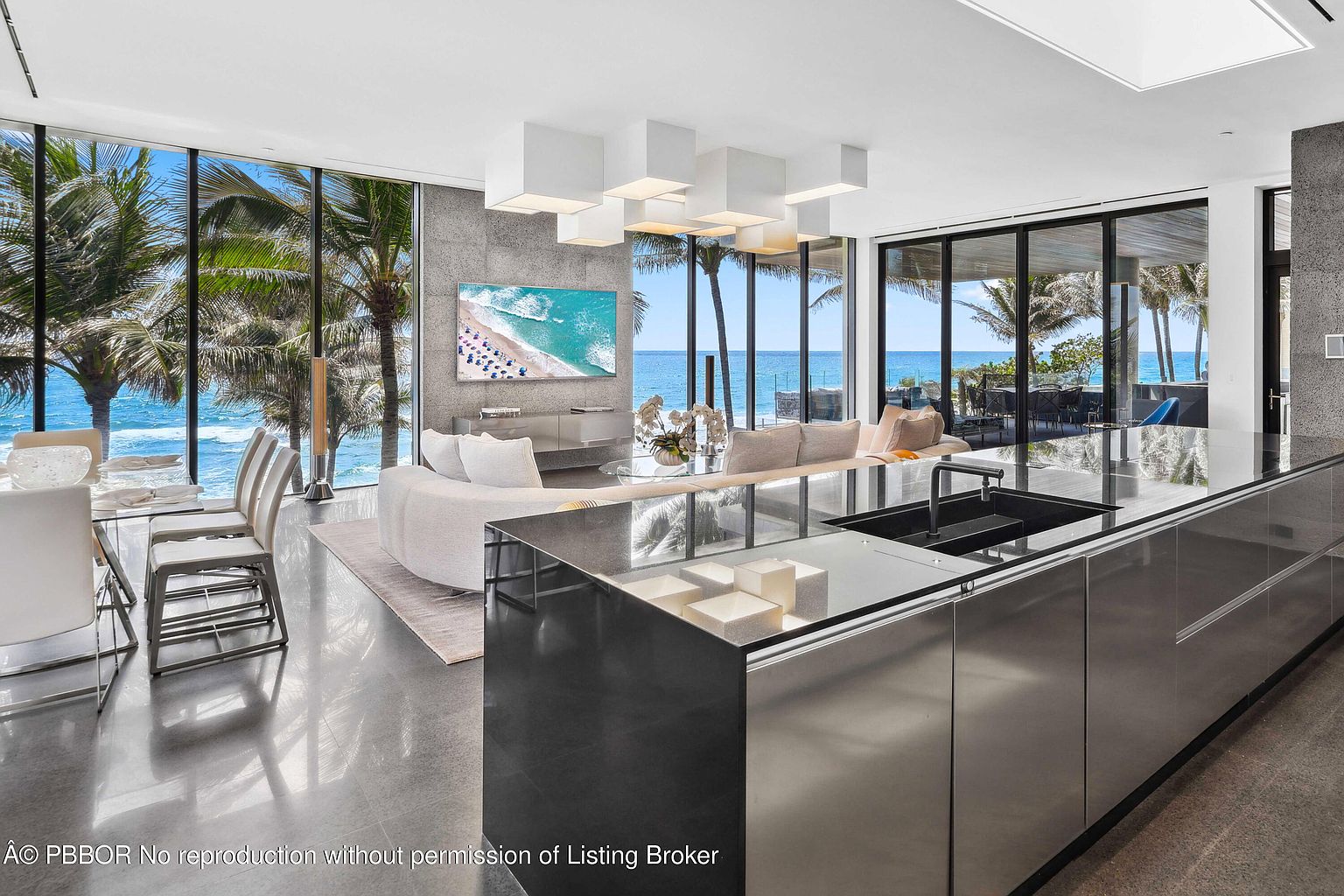 This interior shot showcases a luxurious living space with floor-to-ceiling windows offering stunning ocean views framed by palm trees. The room features a modern design with a sleek kitchen island, a curved sofa, and a contemporary lighting fixture. The overall impression is one of high-end coastal living with an emphasis on natural light and open space.