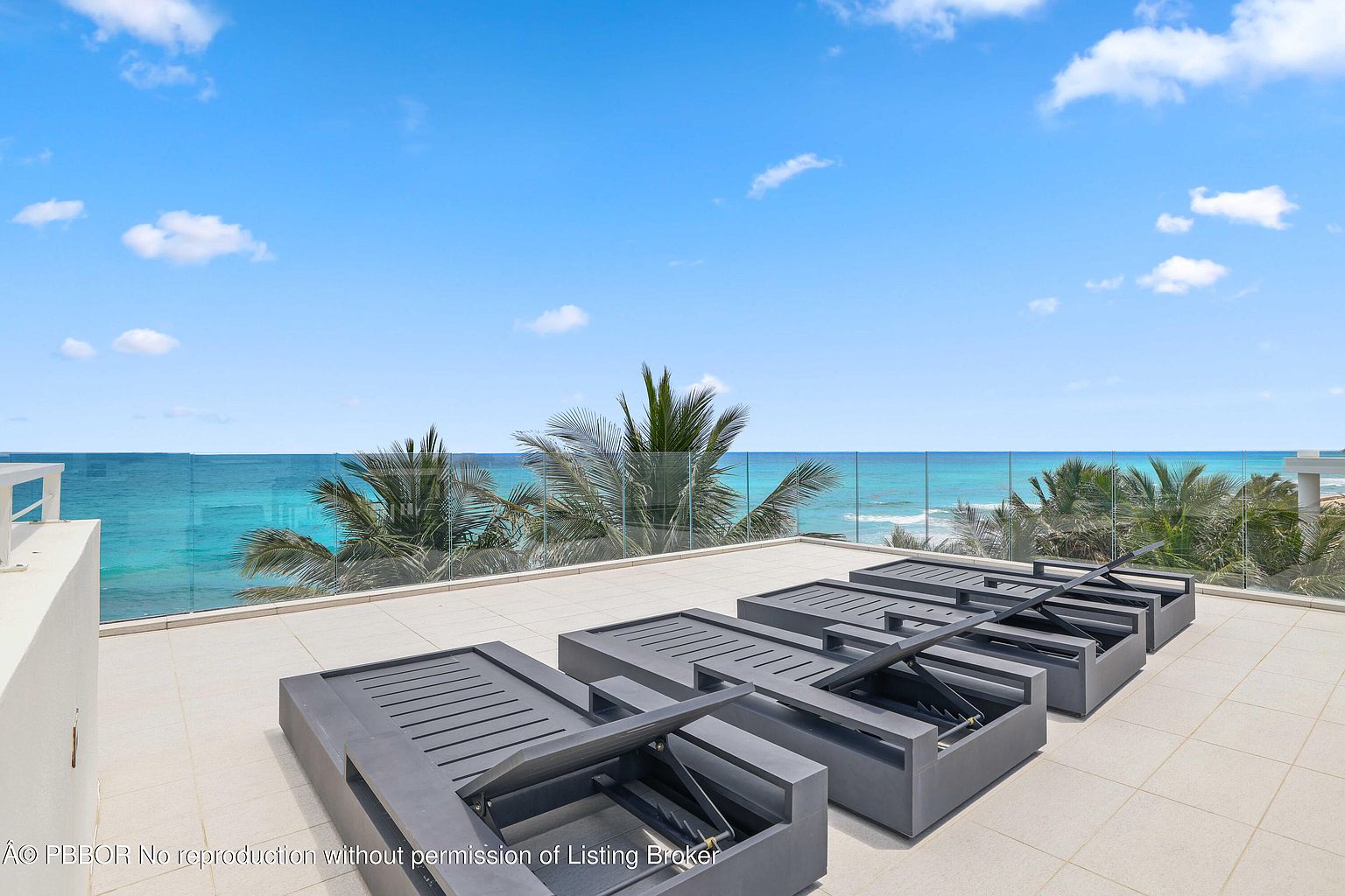 This image showcases a luxurious patio or balcony with stunning ocean views. The space is furnished with modern, dark-toned lounge chairs, arranged neatly on a tiled surface. A glass railing provides an unobstructed view of the turquoise ocean and palm trees, creating a serene and upscale outdoor living area.