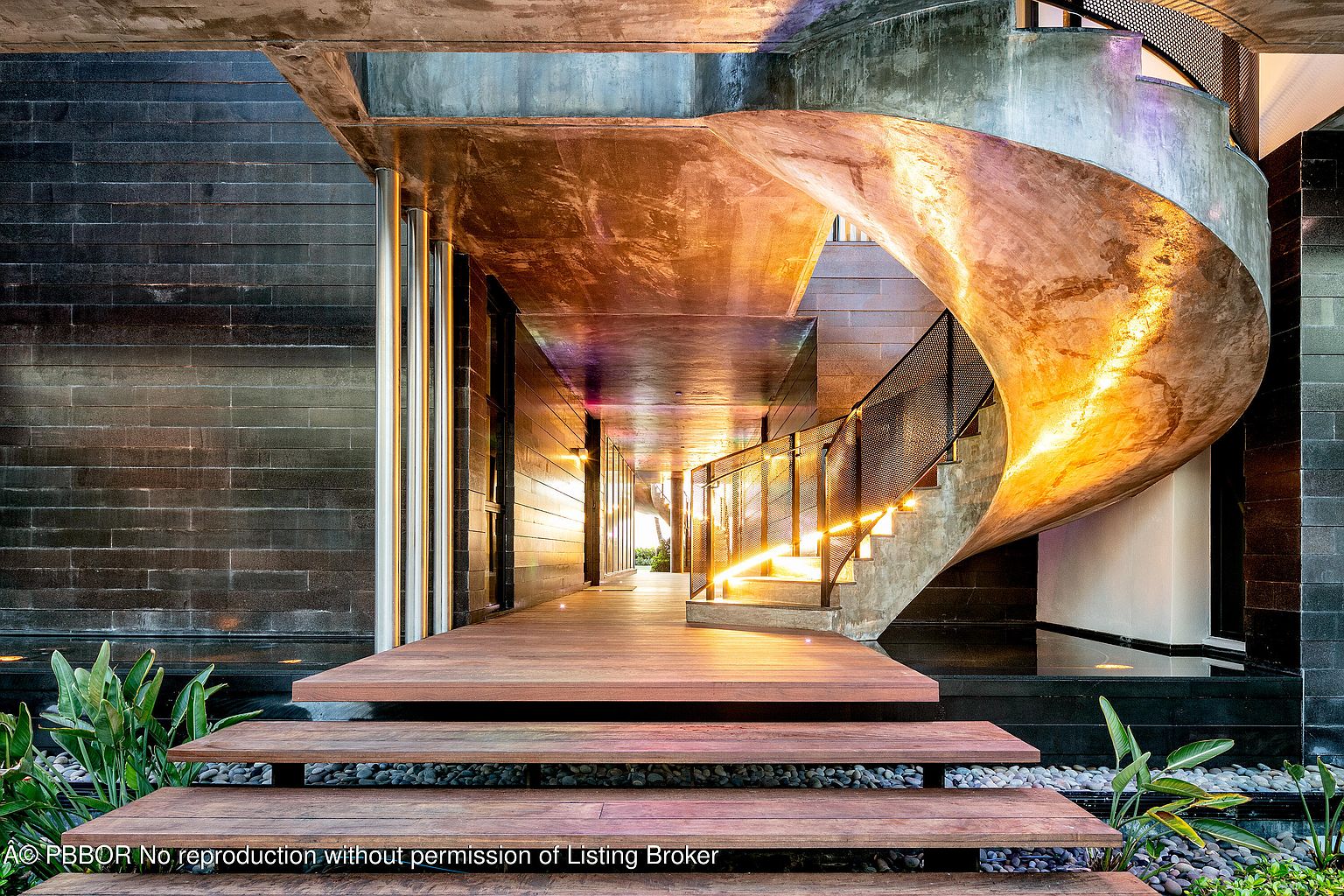 This image showcases a modern home's entryway, featuring a striking spiral staircase and a wooden walkway leading to the entrance. The exterior walls are clad in dark, textured material, contrasting with the warm tones of the wood and the illuminated staircase. The design emphasizes a blend of natural elements and contemporary architecture, creating a sophisticated and inviting atmosphere.