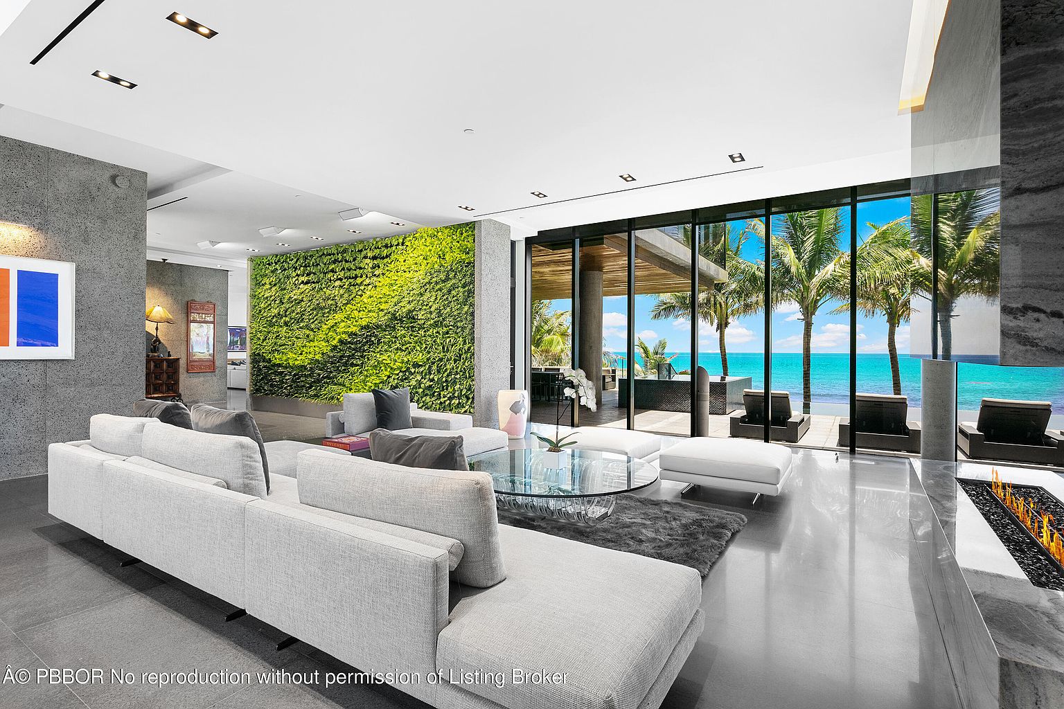 This is a luxurious living room featuring a large sectional sofa, a glass coffee table, and a striking green wall. Floor-to-ceiling windows offer stunning views of the ocean and palm trees, creating a seamless indoor-outdoor living experience. The polished floors and modern fireplace add to the room's sophisticated ambiance.