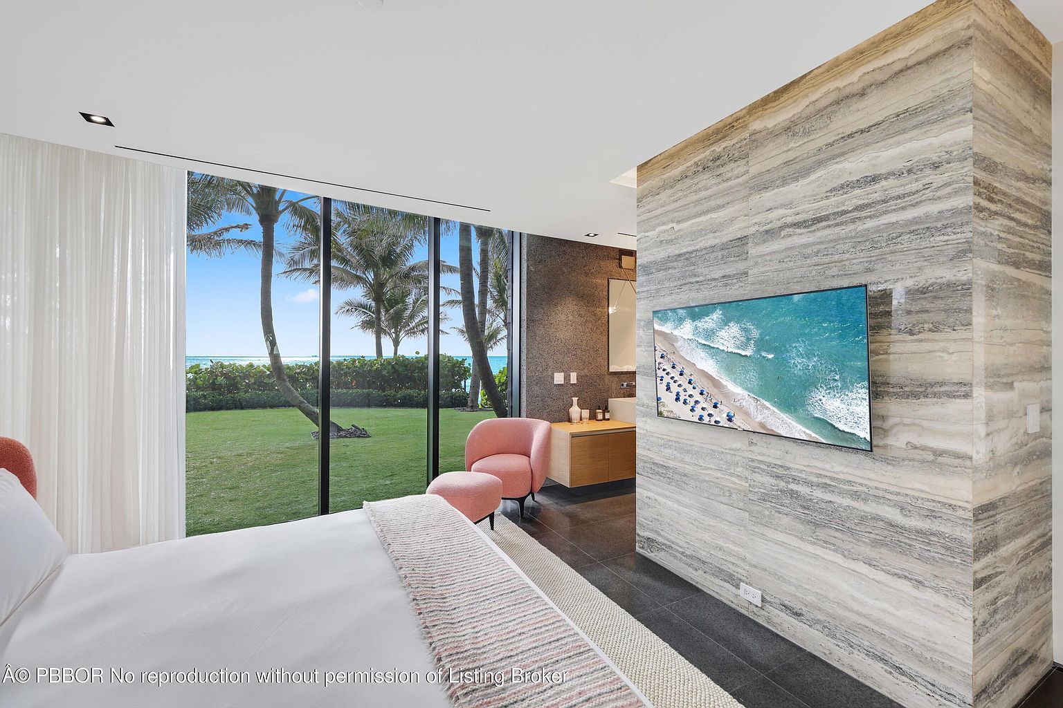 This is a luxurious primary bedroom featuring a large window with a view of palm trees and the ocean. The room has a modern design with a statement wall featuring a television, a pink chair, and a light-colored rug. The bed is neatly made with white linens and a patterned throw, creating a serene and inviting atmosphere.