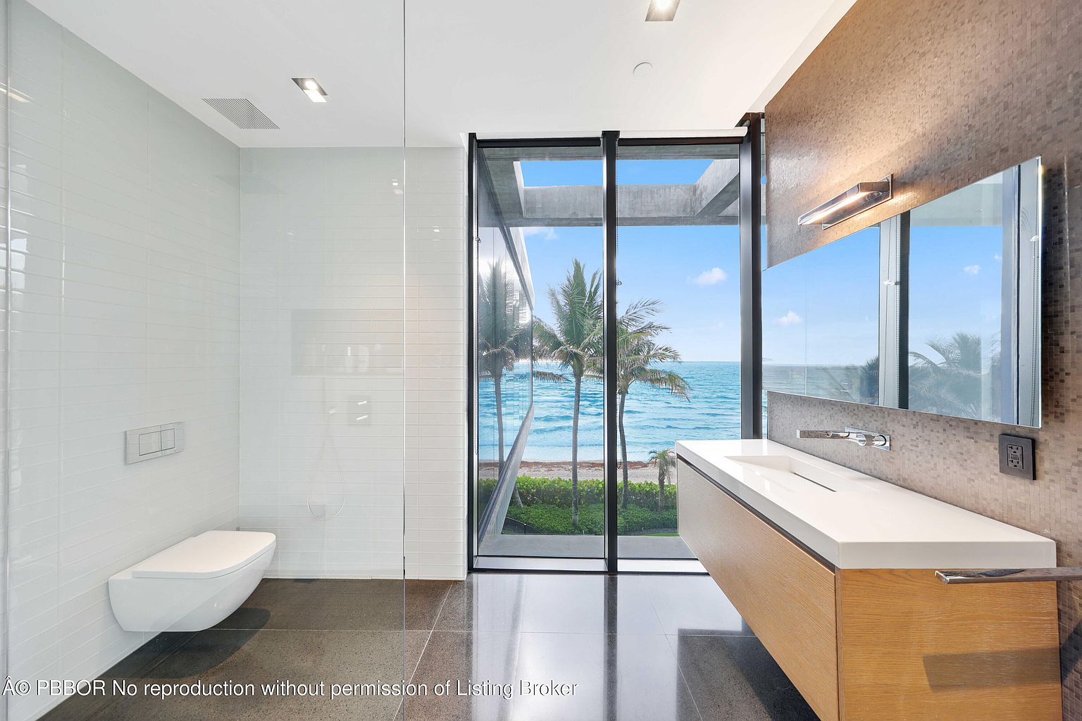 This is a modern primary bathroom featuring a sleek, wall-mounted toilet and a floating vanity with a white countertop and wood-tone base. The shower area is enclosed with glass, and a large window offers a stunning ocean view with palm trees. The walls are a combination of white tile and mosaic tile, creating a luxurious and contemporary space.