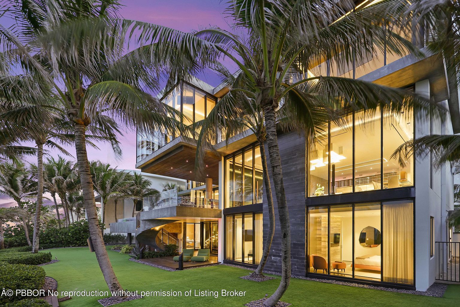 This is a rear view of a modern, multi-story luxury home featuring expansive glass windows and a concrete and stone facade. The property includes a well-manicured lawn, palm trees, and a spiral staircase leading to an upper level patio. The large windows offer views into the interior spaces, showcasing the home's open design and luxurious finishes.