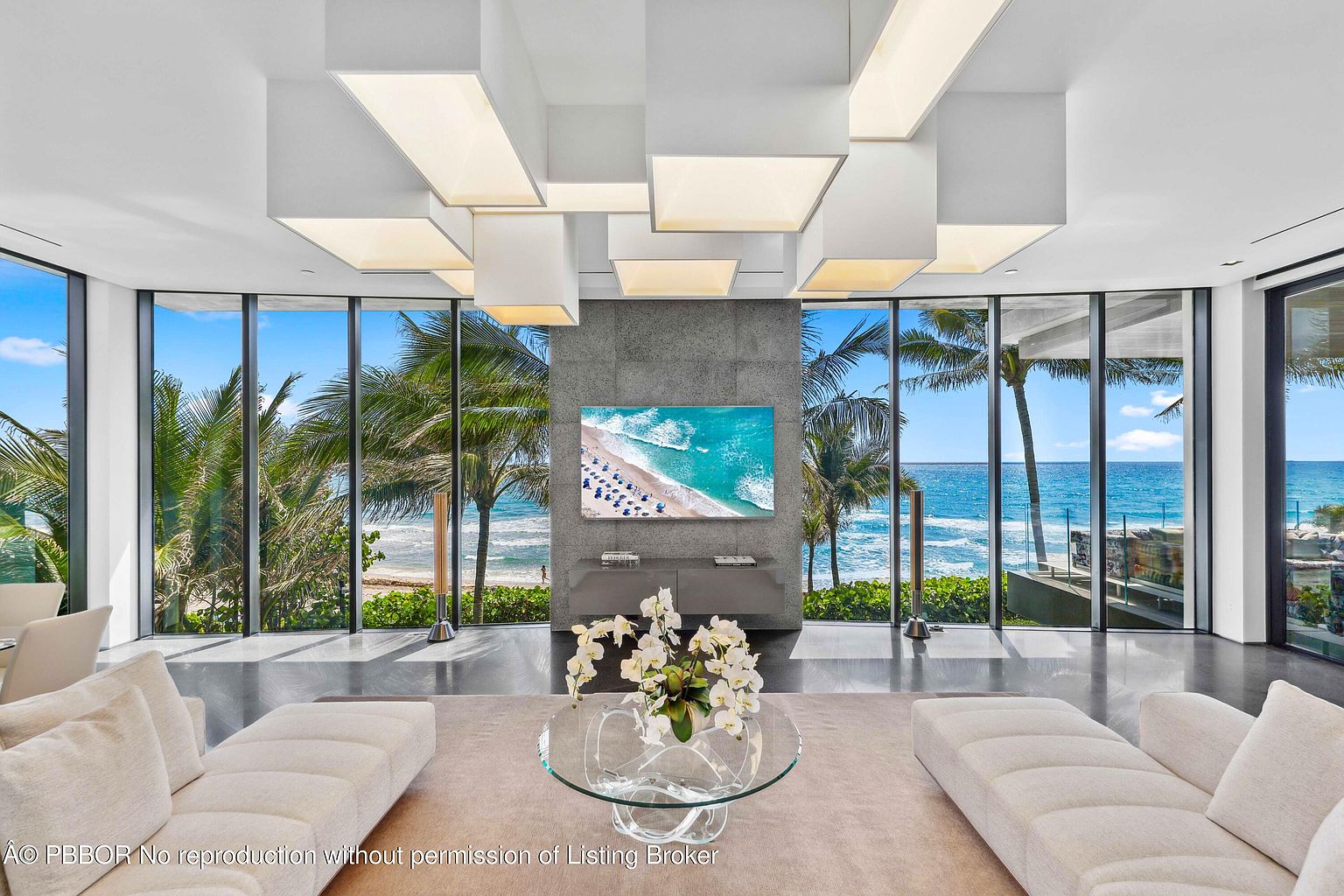 This is a luxurious living room with floor-to-ceiling windows offering stunning ocean views framed by palm trees. The room features modern, minimalist decor with two light-colored sofas, a glass coffee table with floral arrangement, and a large flat-screen TV mounted on a gray accent wall. The unique ceiling lighting fixtures add a contemporary touch to the space.