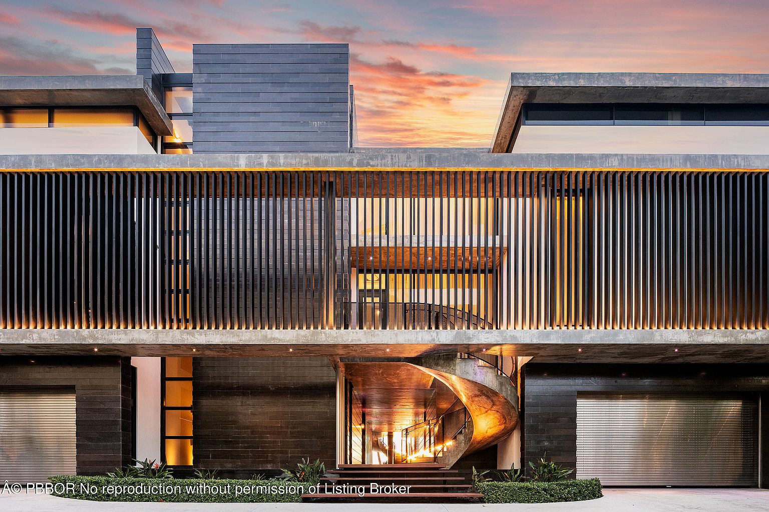 This is a striking front view of a modern architectural masterpiece. The building features a unique facade with vertical slats, a grand spiral staircase leading to the entrance, and a combination of concrete and dark materials. The warm lighting contrasts beautifully with the dramatic sunset sky, creating an inviting and luxurious ambiance.