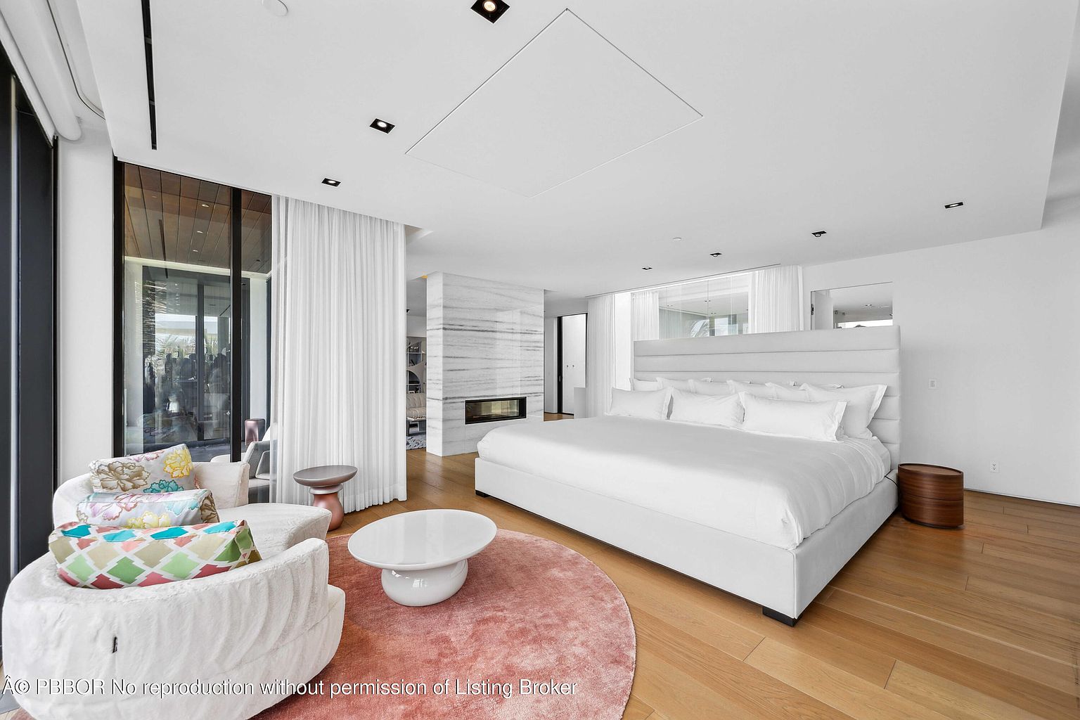 This is a bright and spacious primary bedroom featuring a large white platform bed with a padded headboard, complemented by a round pink rug and modern white furniture. The room boasts hardwood floors, a sleek fireplace with marble surround, and large windows with sheer curtains, creating a luxurious and serene atmosphere. The perspective is from the corner of the room, capturing the full breadth of the space.