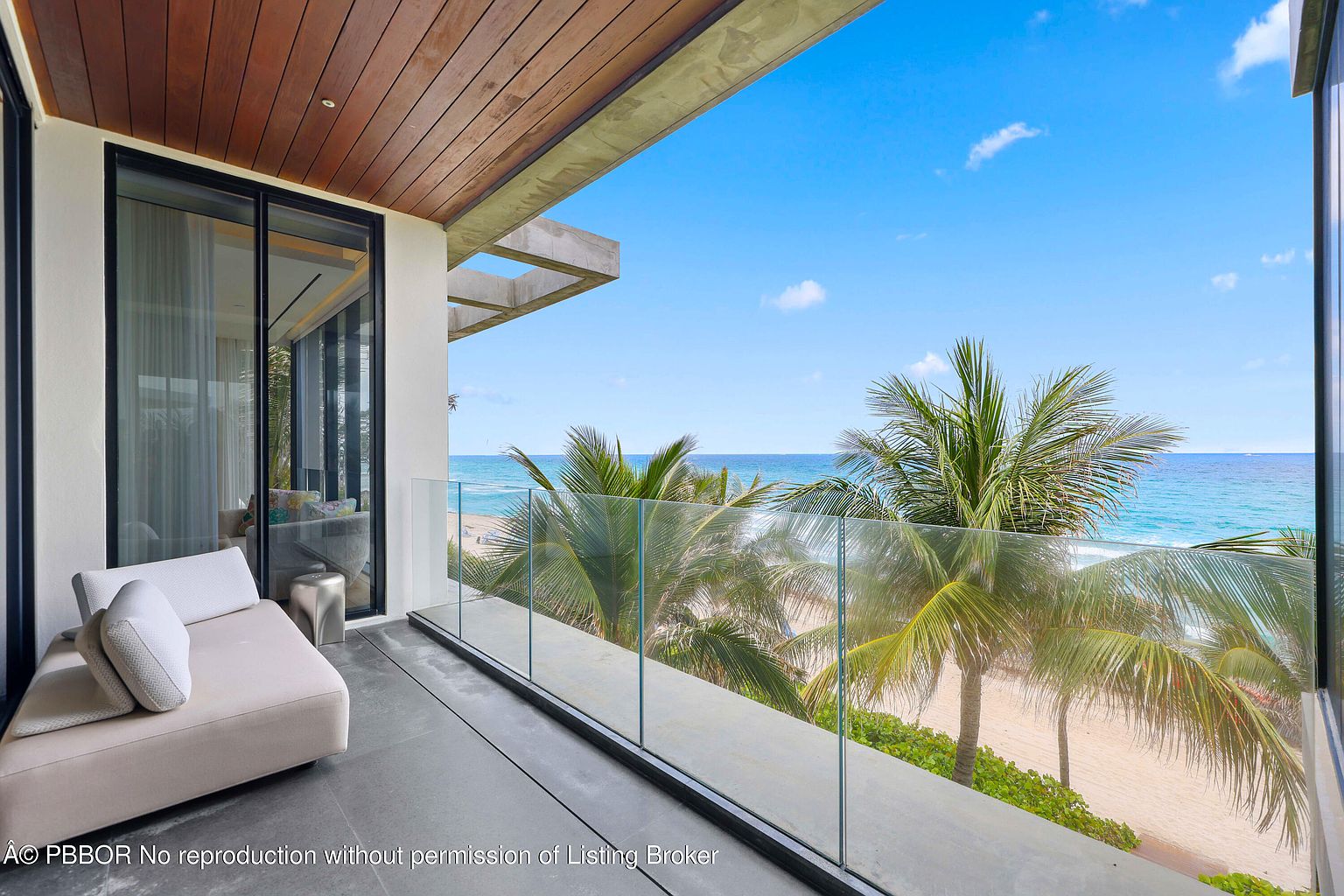 This image showcases a luxurious balcony with a modern design, featuring a sleek lounge chair and a glass railing that offers unobstructed views of the ocean and palm trees. The balcony has a gray stone floor and a wood-paneled ceiling, adding warmth to the contemporary aesthetic. The scene evokes a sense of relaxation and high-end coastal living.