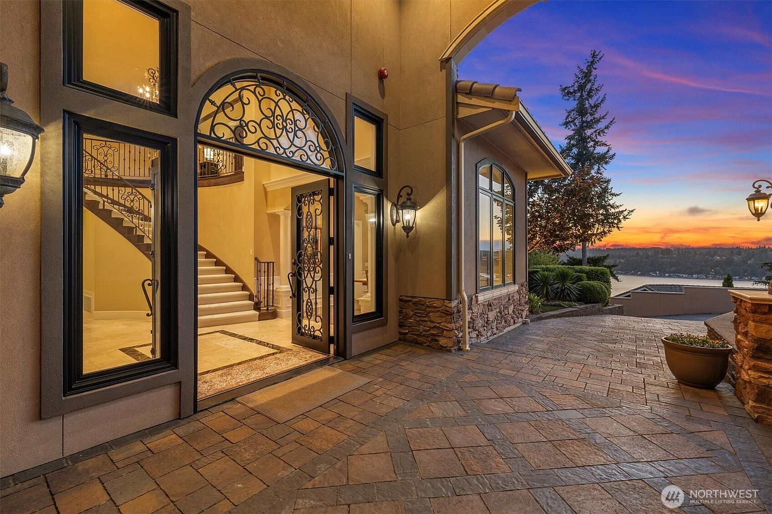This is an inviting entryway to a luxurious home, featuring an ornate wrought-iron door and sidelights that lead into a grand foyer with a staircase. The exterior is finished with stone accents and a paved driveway, while the warm lighting and sunset in the background create a welcoming ambiance. The architecture suggests high-end craftsmanship and attention to detail.