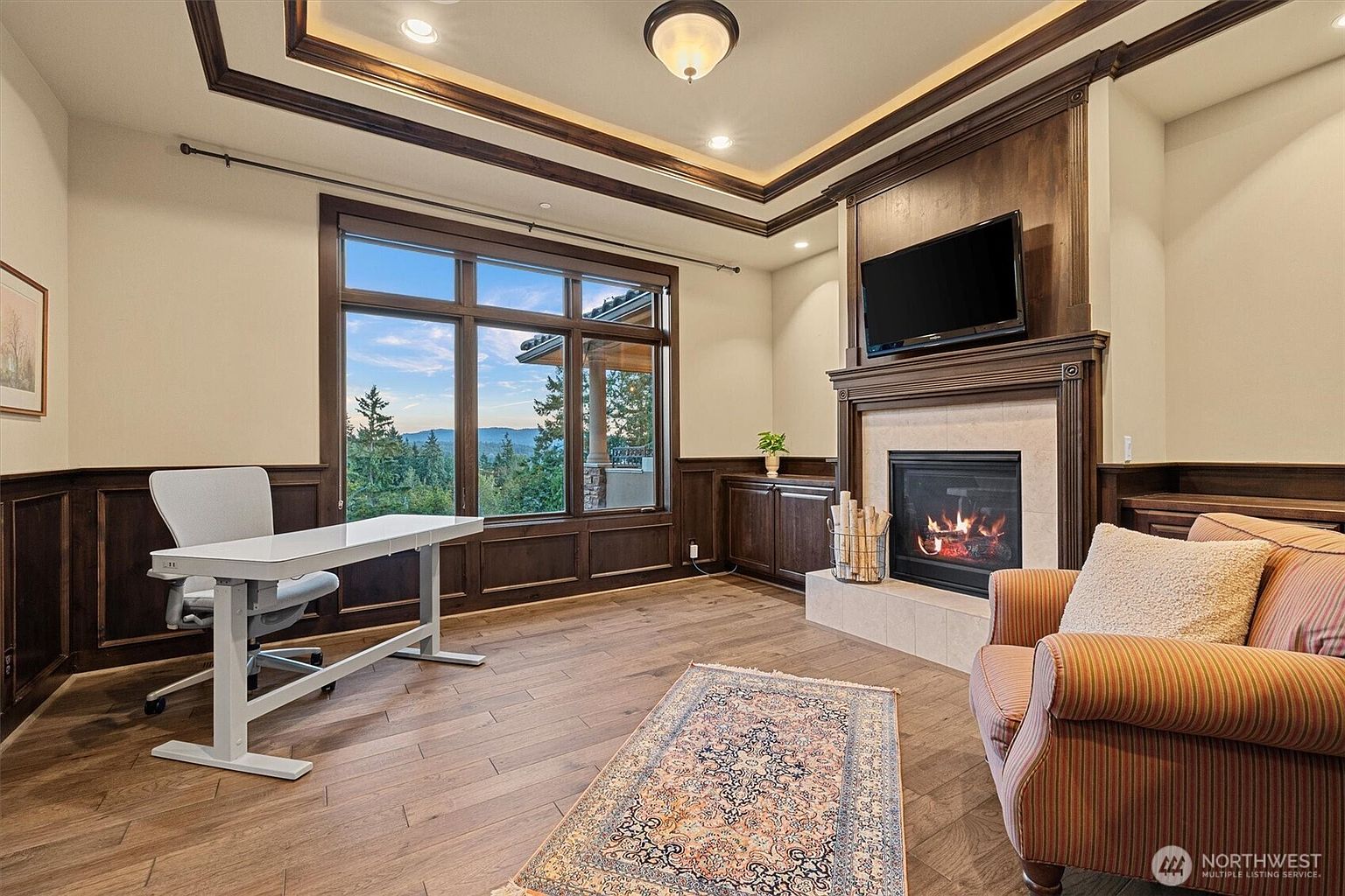 This is an interior shot of a well-appointed office or study. The room features dark wood paneling, a large window with a scenic view, a modern white desk, and a comfortable armchair next to a fireplace with a mounted television above. The space is designed for both work and relaxation, offering a blend of functionality and comfort.
