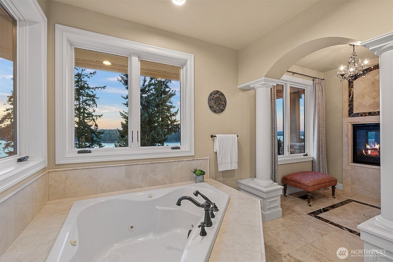 This is a luxurious primary bathroom featuring a large soaking tub positioned beneath a window with a scenic view. The room is decorated with neutral tones, marble accents, and elegant fixtures, including a chandelier and a fireplace, creating a spa-like atmosphere. The design emphasizes relaxation and comfort.