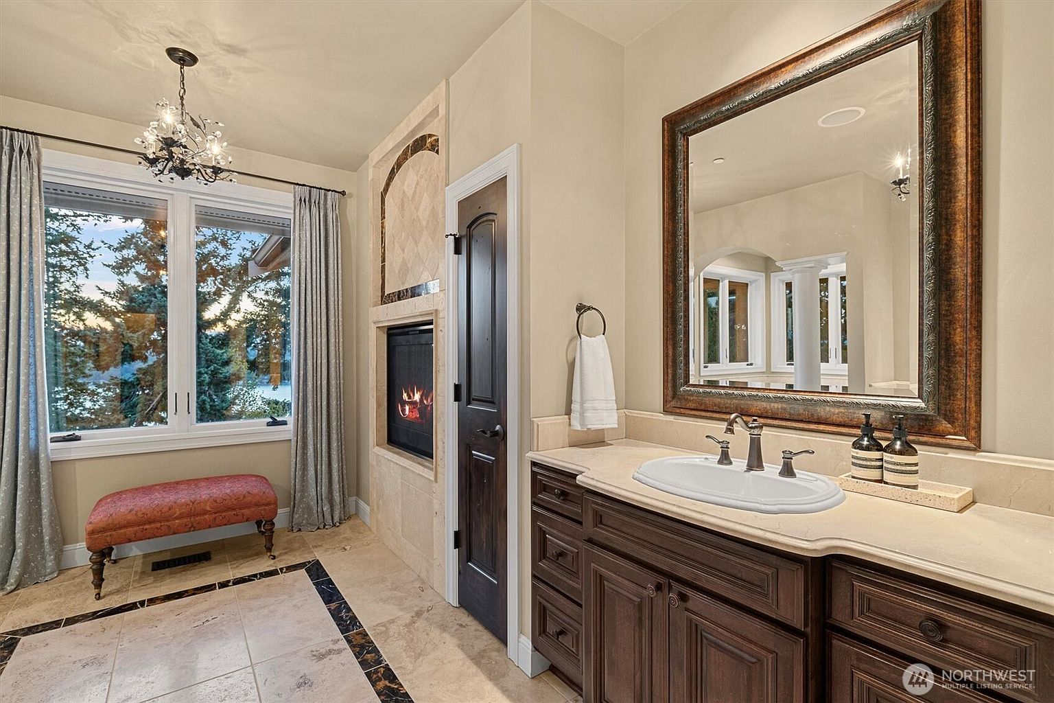 This is a luxurious primary bathroom featuring a dark wood vanity with a light countertop and an oval sink. A large framed mirror hangs above the vanity. The bathroom also includes a fireplace, a window with a view, and a decorative chandelier, creating a spa-like atmosphere.
