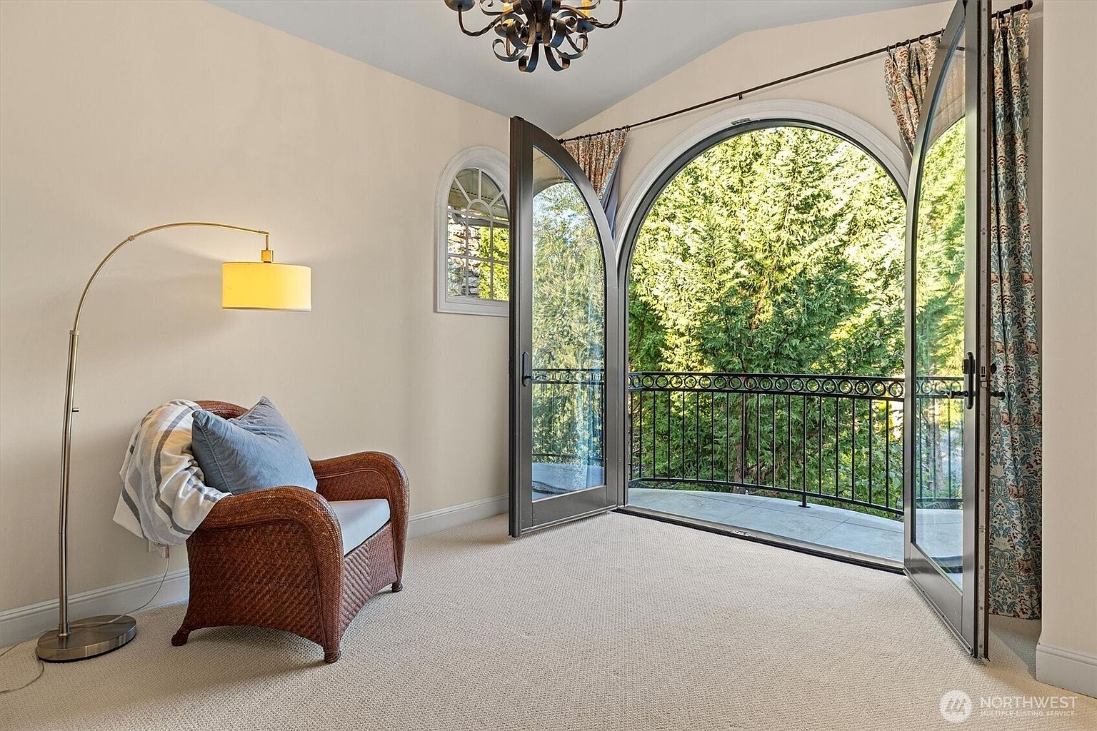 This is an interior shot of a bedroom featuring a cozy reading nook. The room is well-lit, with natural light streaming in through arched glass doors that lead to a balcony. A comfortable wicker chair with a blue pillow sits next to an arched floor lamp, creating an inviting space for relaxation.