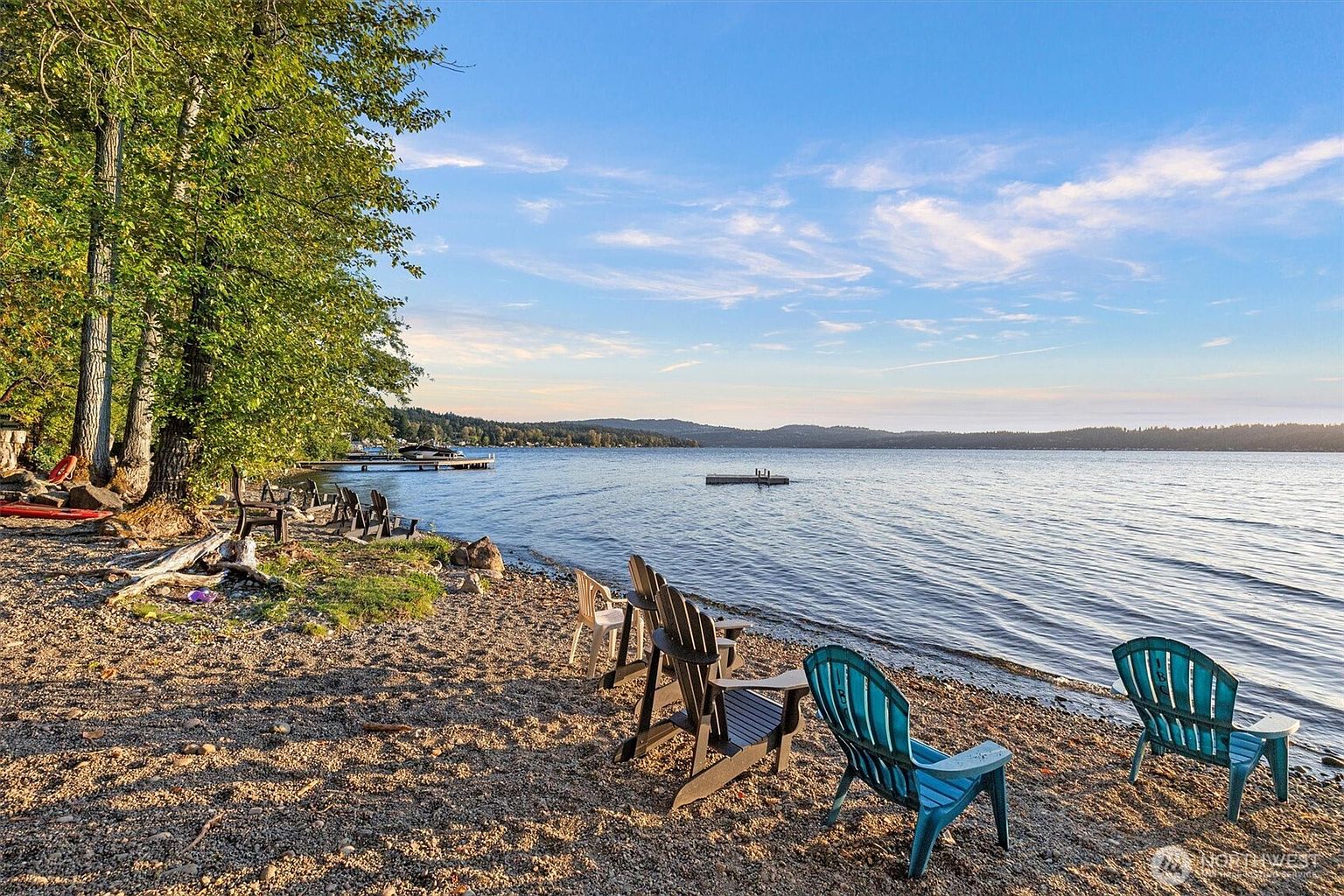 This serene lakeside scene showcases a sandy beach with several Adirondack chairs arranged for relaxation. The calm water stretches to a distant shoreline with forested hills, under a sky with scattered clouds. The image evokes a sense of tranquility and outdoor living, highlighting the property's access to waterfront amenities.