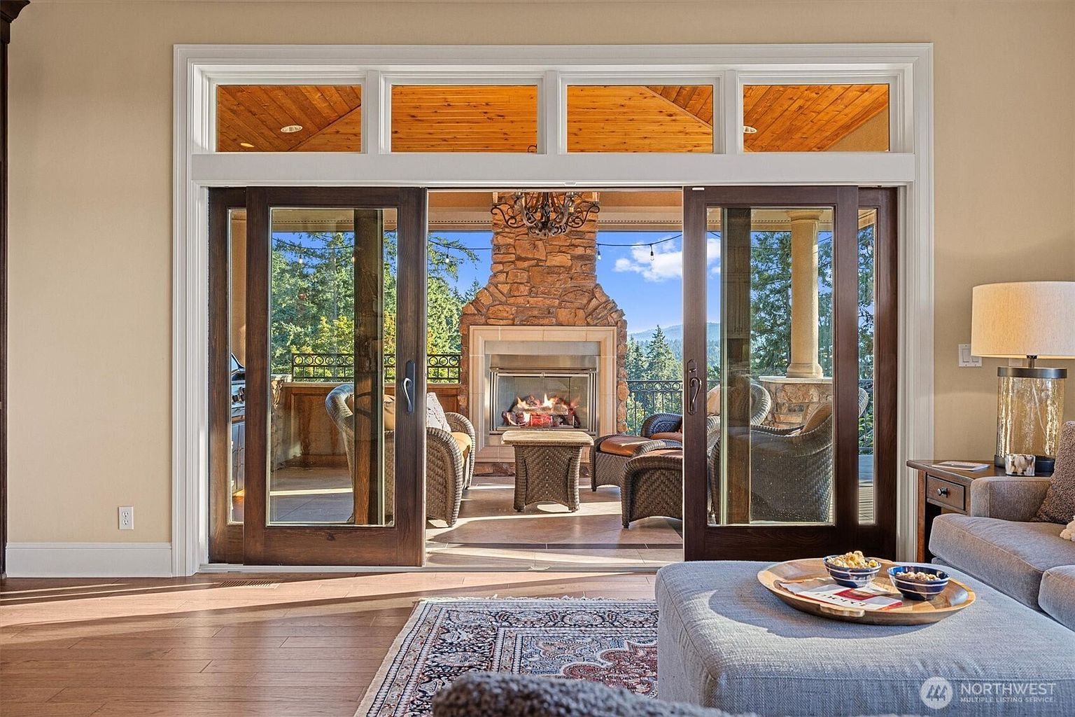 This inviting living room seamlessly blends indoor and outdoor living with large sliding doors opening onto a patio featuring a stone fireplace. The interior boasts hardwood floors, a comfortable seating area with a gray ottoman, and a glimpse of a well-appointed side table with a lamp. The overall impression is one of relaxed elegance and connection to nature.