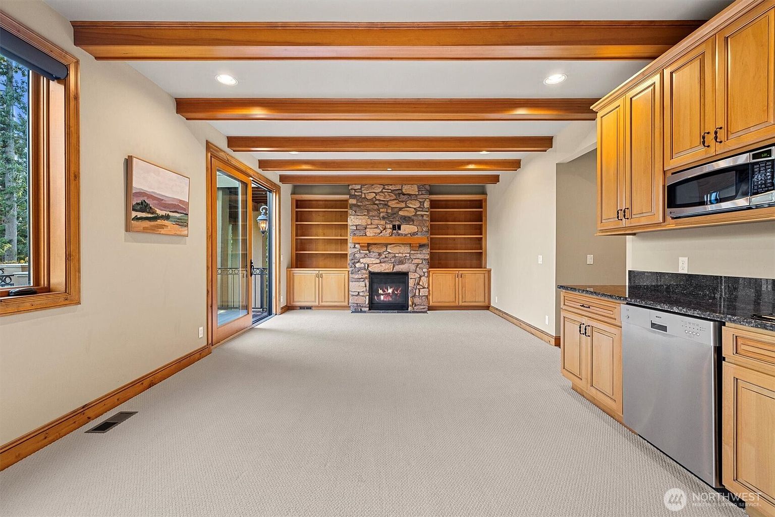This interior shot showcases a spacious living room featuring a stone fireplace flanked by built-in bookshelves, creating a cozy focal point. The room is enhanced by wooden ceiling beams and trim, adding warmth and character. A sliding glass door provides access to the outside, while a partial view of the kitchen area with wooden cabinets and stainless steel appliances is visible on the right.