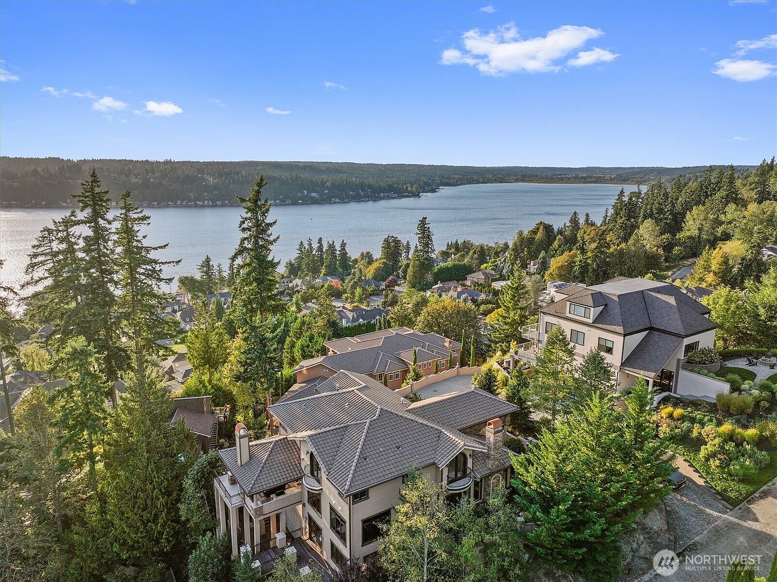 This aerial view showcases a luxurious multi-story home with a tile roof, multiple balconies, and manicured landscaping. The property is nestled among mature trees, offering privacy and scenic views of the lake in the background. The overall impression is one of elegance and exclusivity.