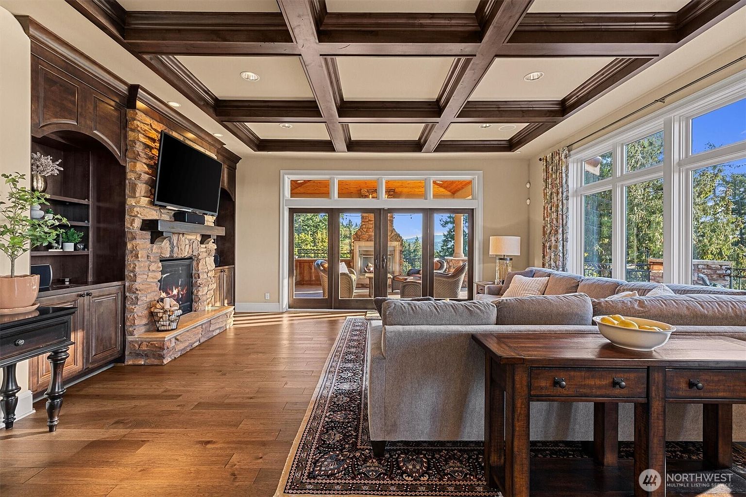 This is an interior shot of a luxurious living room featuring a coffered ceiling with dark wood beams, a stone fireplace with a built-in entertainment center, and large windows offering natural light. The room is furnished with a comfortable gray sectional sofa and a wooden side table, creating a warm and inviting atmosphere. The perspective is from the entrance of the room, showcasing the depth and layout.