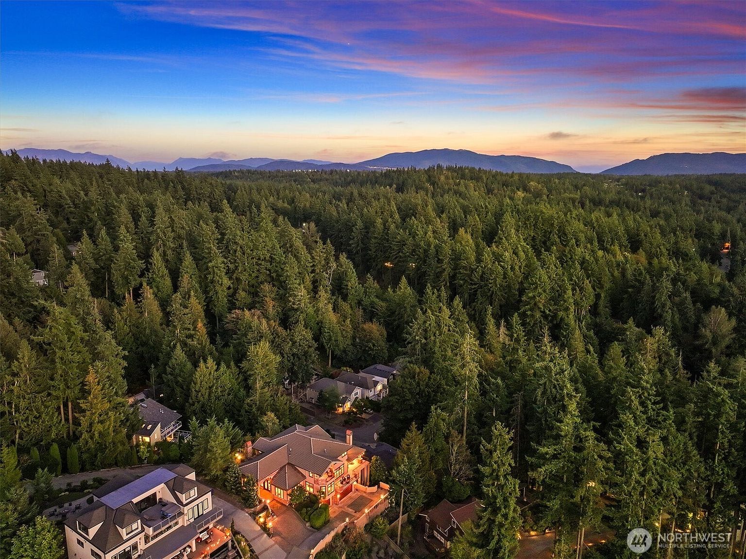 This aerial view showcases a luxurious property nestled among dense evergreen trees, offering a sense of privacy and seclusion. The homes feature elegant architectural details, including well-manicured landscaping and illuminated exteriors. The backdrop of distant mountains under a vibrant sunset sky enhances the property's appeal, suggesting a serene and upscale living environment.