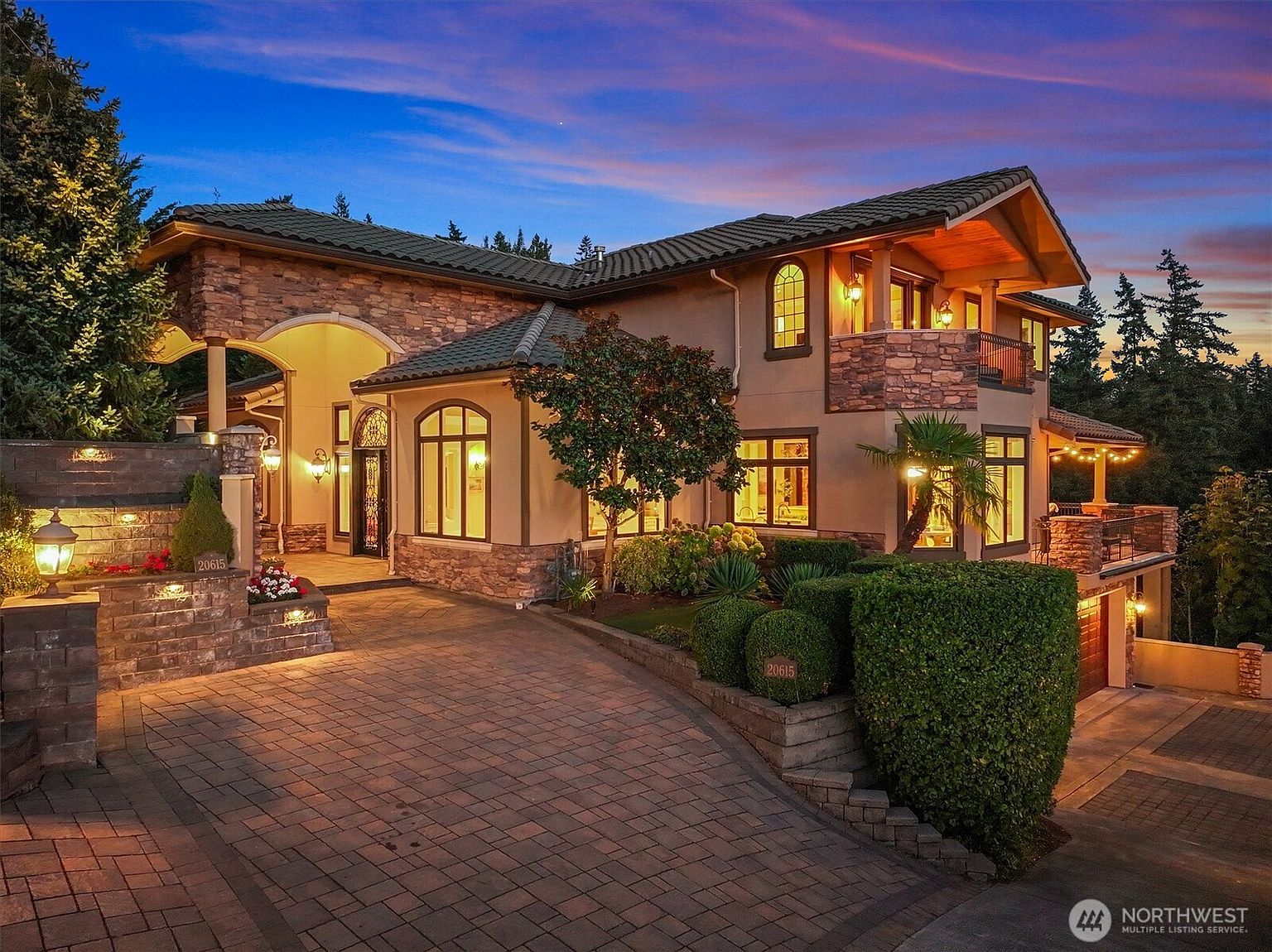 This is a beautifully illuminated front view of a luxurious two-story home at dusk. The house features a combination of stone and stucco exterior, arched entryway, and a well-manicured landscape. The brick driveway leads up to the house, enhancing its curb appeal and creating a welcoming atmosphere.