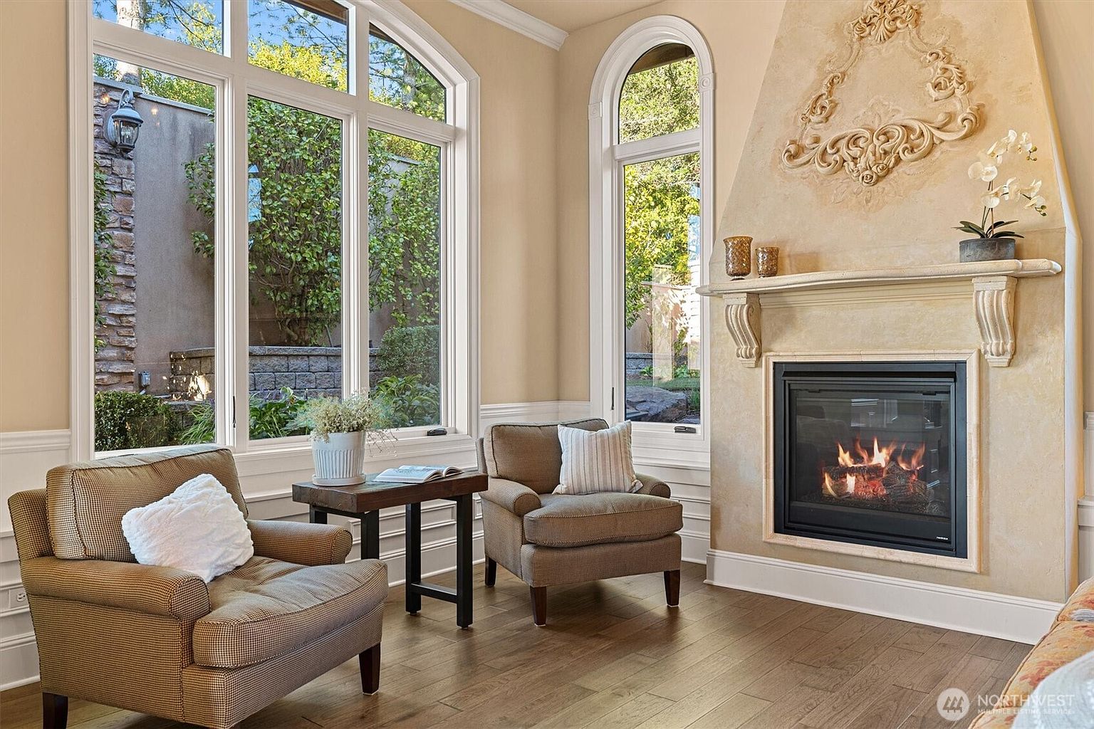 This inviting living room features two comfortable armchairs flanking a small side table, with large windows offering views of the lush greenery outside. A fireplace with an ornate mantel adds a touch of elegance, while the hardwood floors contribute to the room's warm and cozy atmosphere. The room is well-lit and appears to be a comfortable space for relaxation.