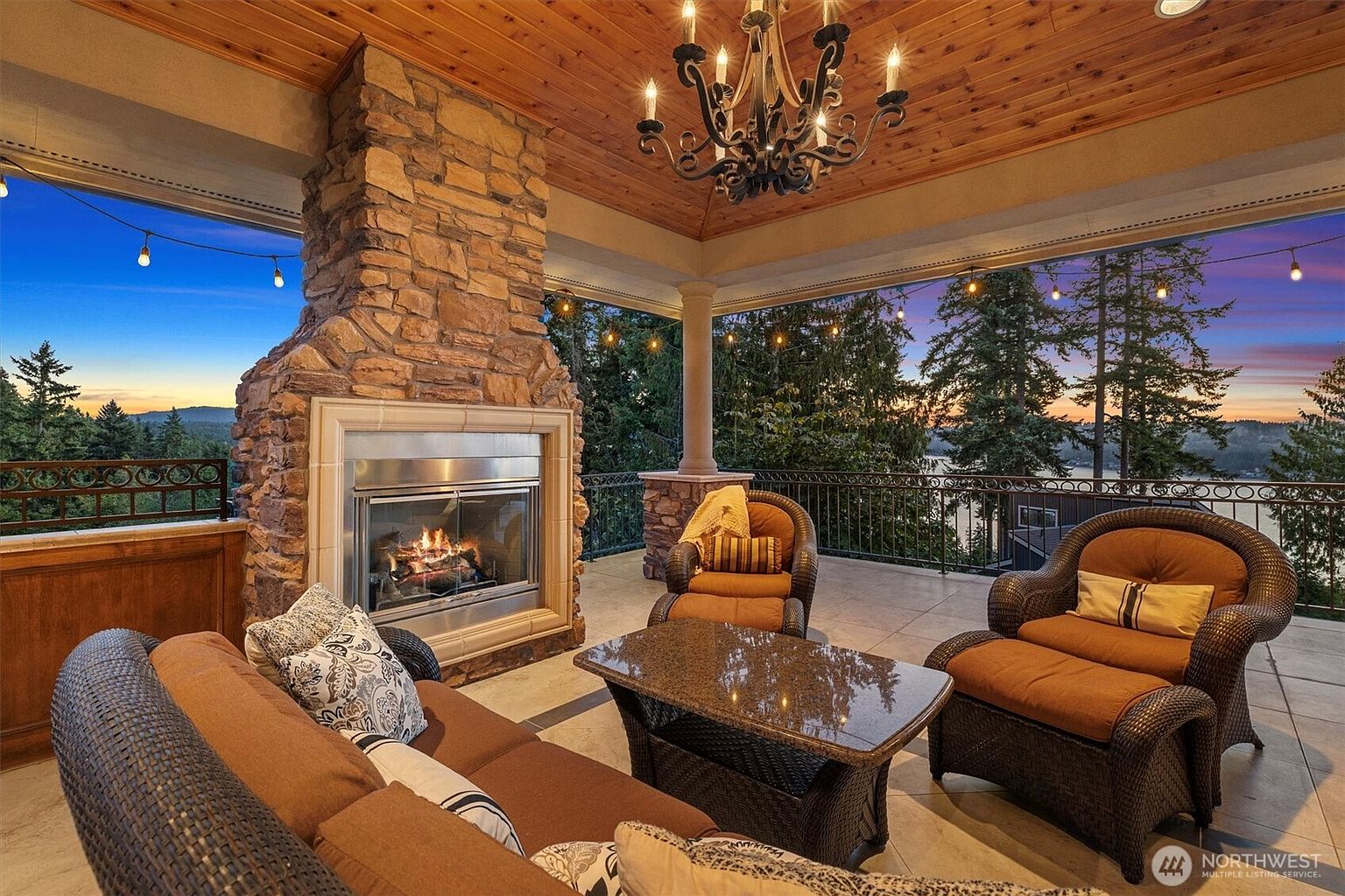 This is a luxurious outdoor patio area featuring comfortable wicker furniture, including a sofa and armchairs, arranged around a stone-topped coffee table. A grand stone fireplace adds warmth and ambiance, while a decorative chandelier hangs from the wooden ceiling. The patio offers stunning views of the surrounding landscape and a lake at sunset, framed by a wrought iron railing.