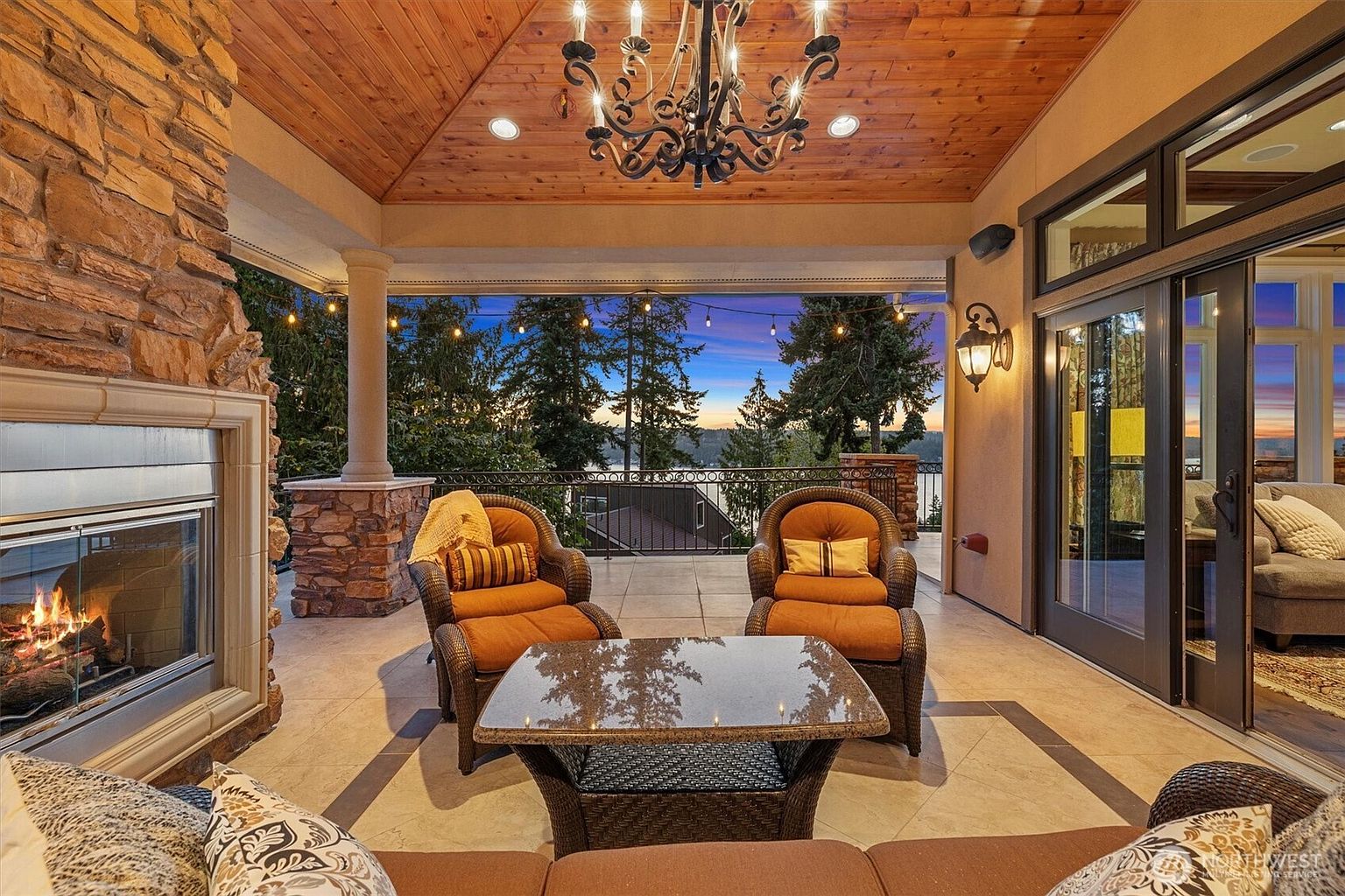 This is a beautifully designed outdoor patio area featuring a stone fireplace, comfortable wicker seating, and a glass-topped table. The space is covered with a wooden ceiling and illuminated by a chandelier, creating a cozy and inviting atmosphere. The patio offers a view of the surrounding trees and a glimpse of the water, enhancing the property's appeal for relaxation and entertainment.